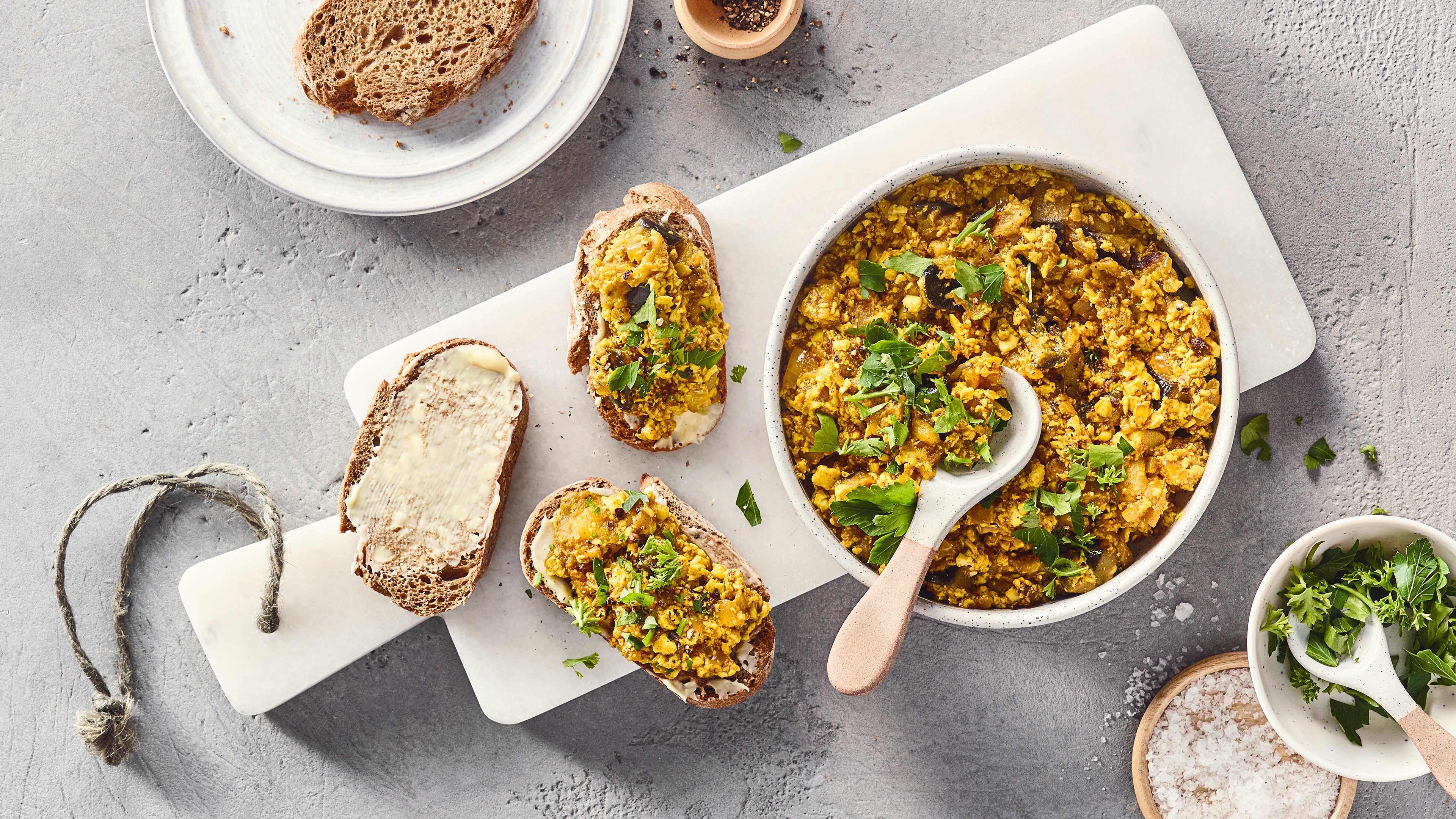 Eine Schüssel mit gelber krümeliger Masse, grünen Kräutern und dunklen Würfeln, daneben Brotscheiben.