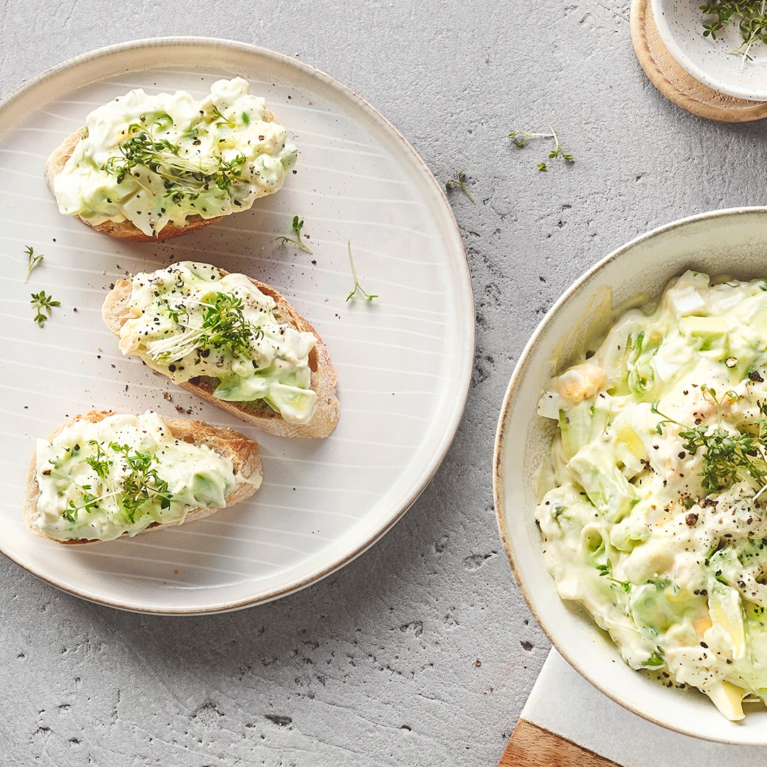 Ein Teller mit drei Brotscheiben, belegt mit weißer Creme, grünen Avocadostücken, schwarzem Pfeffer und frischer Kresse.