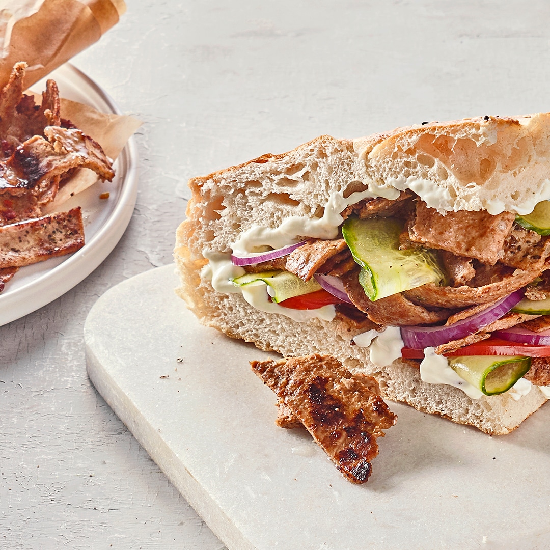 Ein Brett mit aufgeschnittenem Fladenbrot, gefüllt mit gegrilltem Fleisch, Gurken, Tomaten, roten Zwiebeln & weißer Soße.