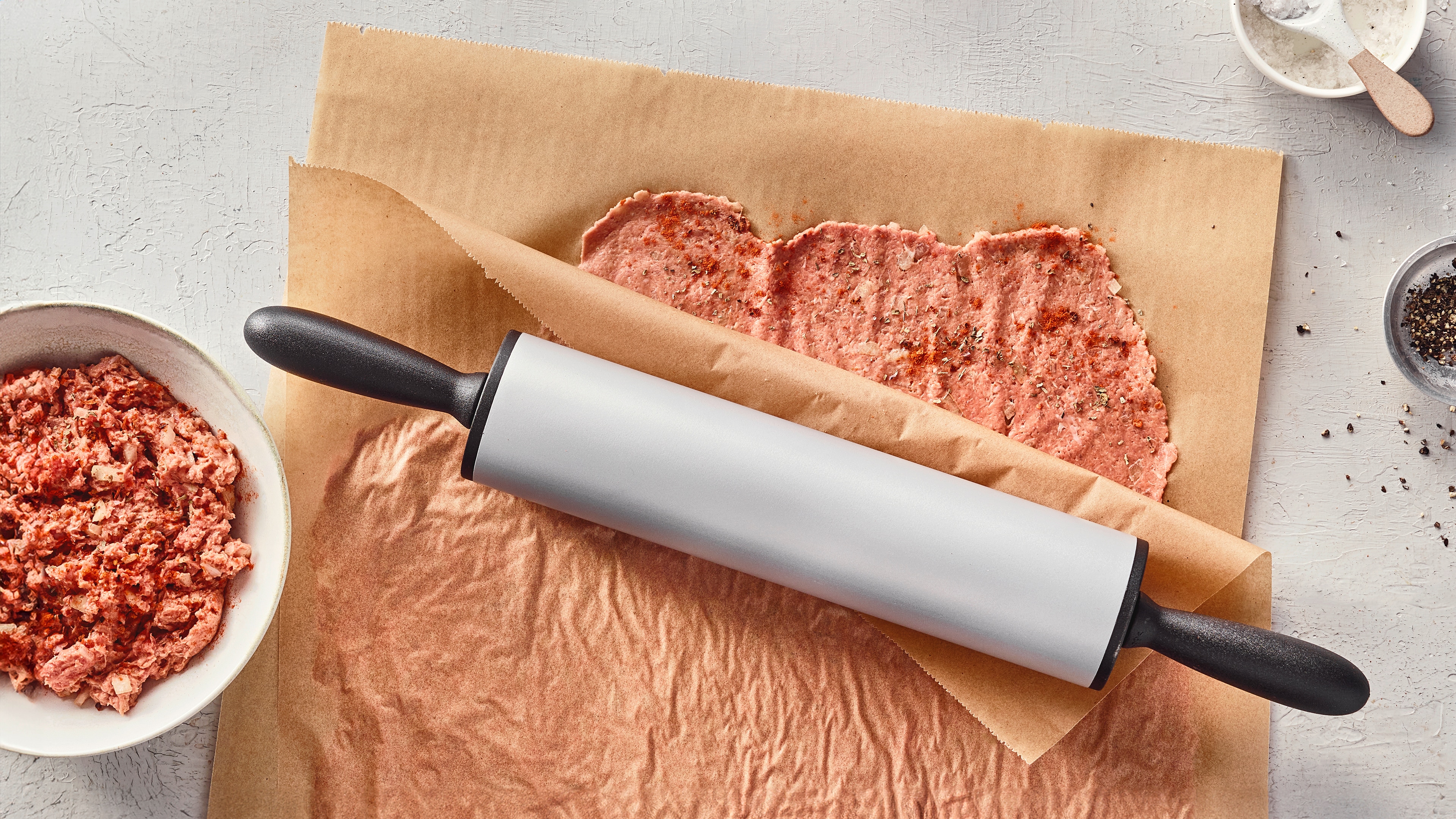 Ein Brett mit ausgerolltem Hackfleisch auf braunem Papier, bestreut mit Gewürzen, daneben eine Schüssel mit Hackfleisch.