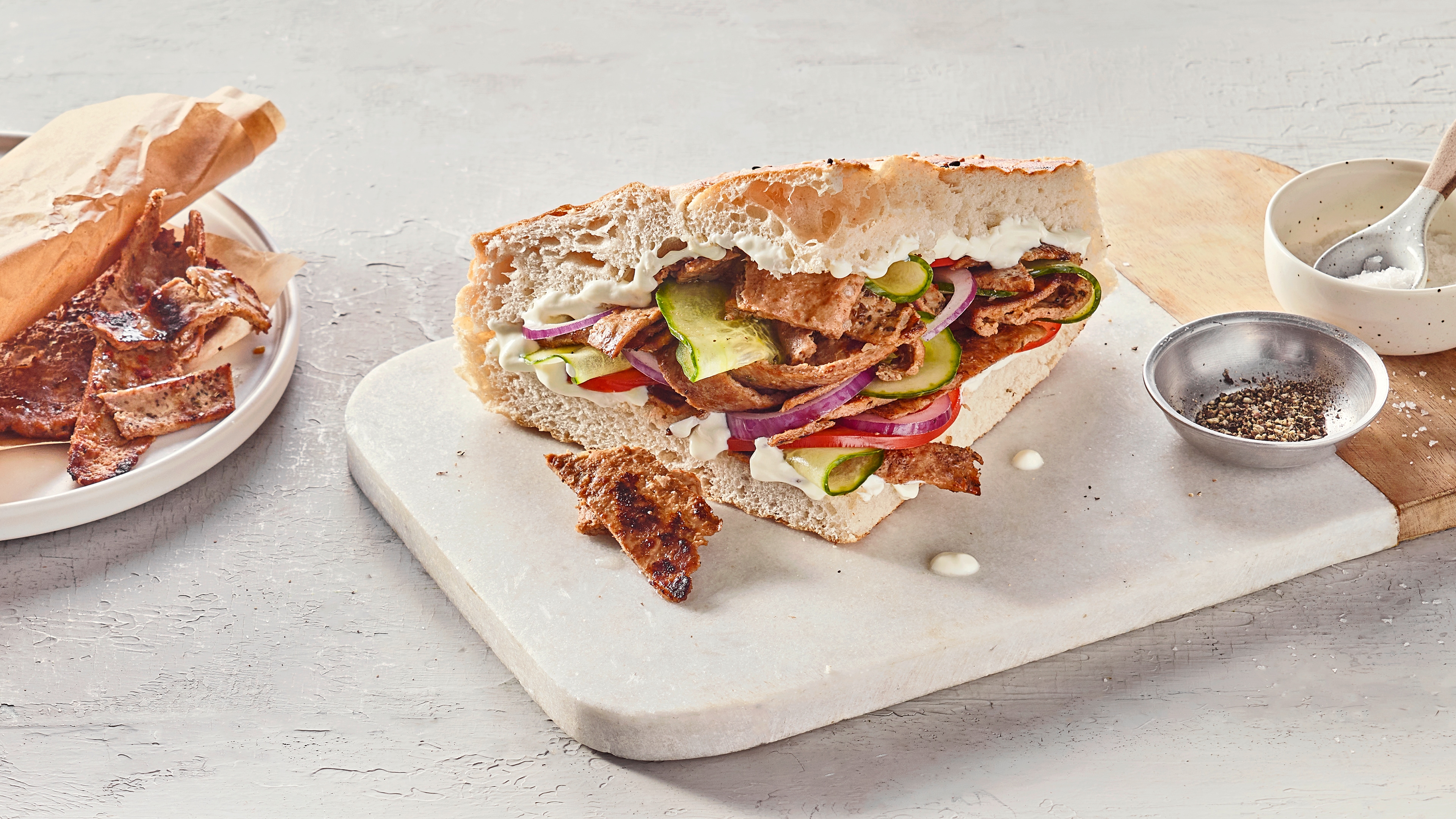 Ein Brett mit aufgeschnittenem Fladenbrot, gefüllt mit gegrilltem Fleisch, Gurken, Tomaten, roten Zwiebeln & weißer Soße.