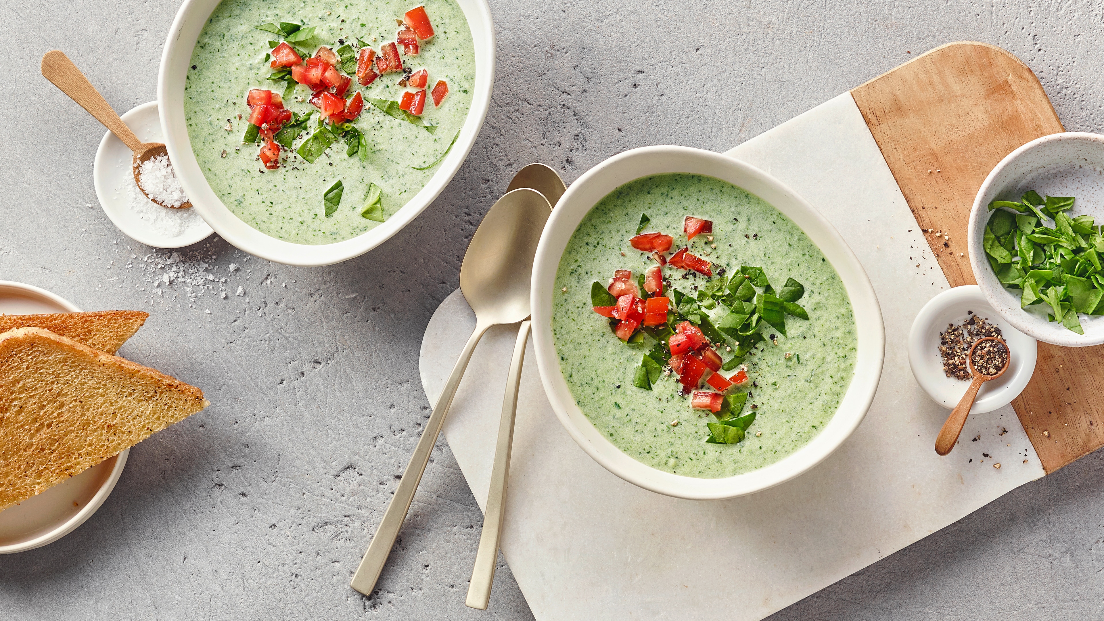 Schalen mit grünlicher cremiger Suppe auf Holzbrett, daneben Toastscheiben.