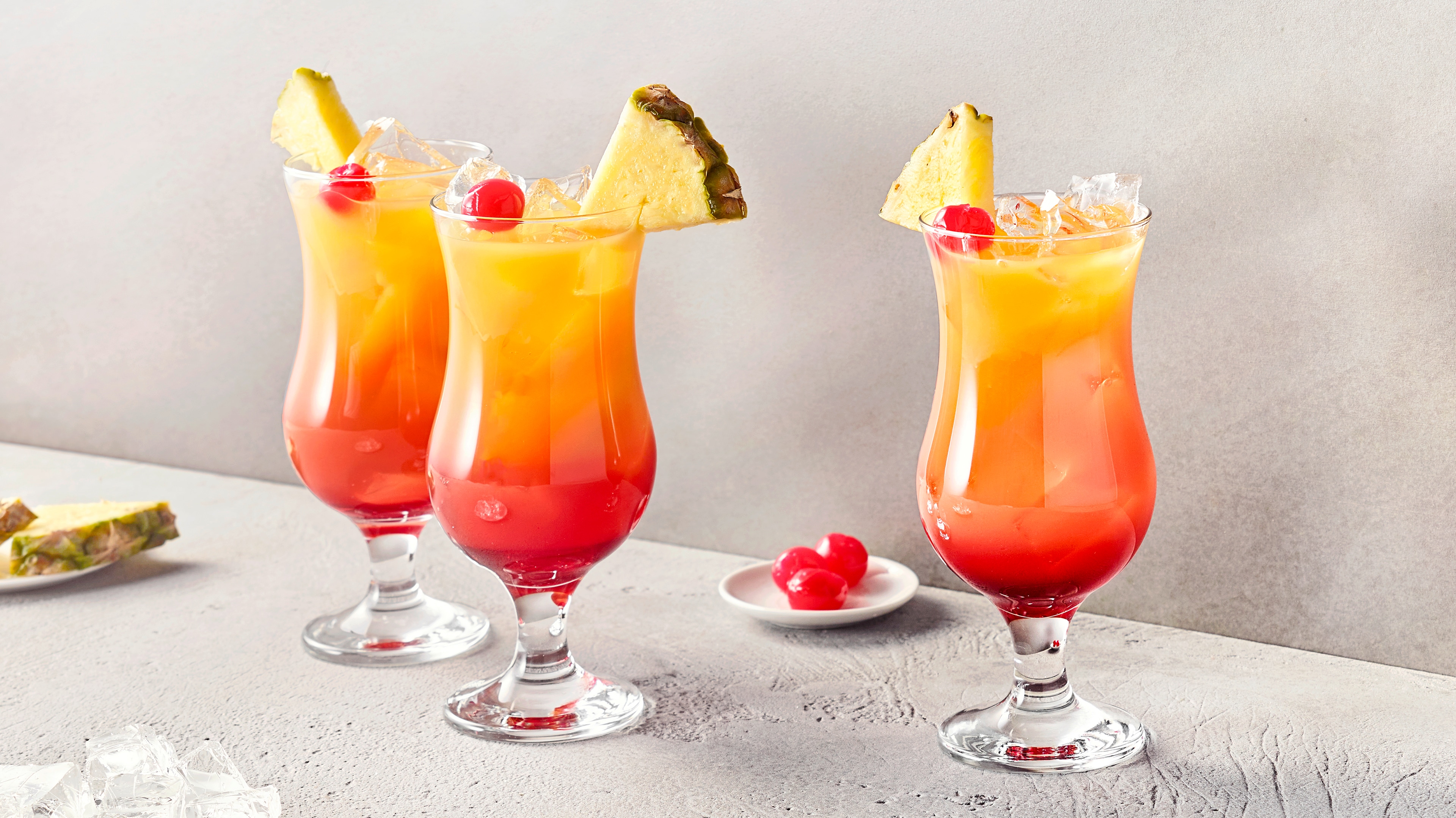 Drei Gläser mit rot-gelber Flüssigkeit, Eiswürfeln, Kirsche und jeweils einem Ananasstück