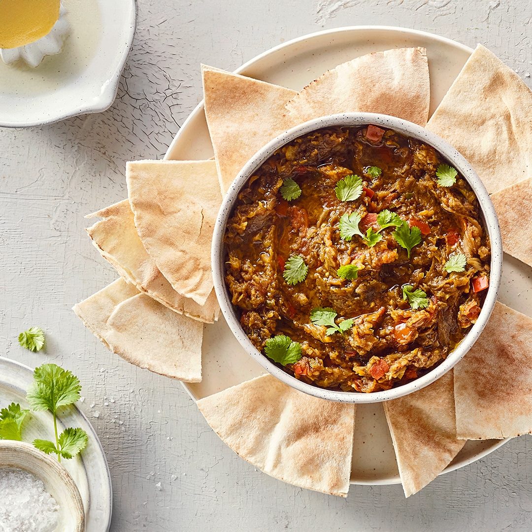 Ein Teller mit hellen, dreieckigen Fladenbrotstücken rund um eine Schale brauner Paste mit grünen Kräutern und kleinen roten Stücken.