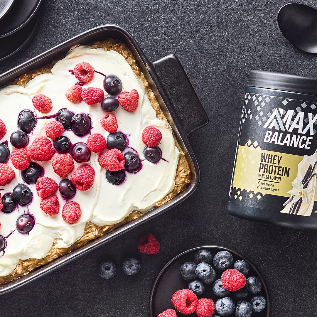 Auflaufform mit gebackenem Teig, heller Creme und Beeren, daneben eine Dose MaxBalance Proteinpulver Vanille