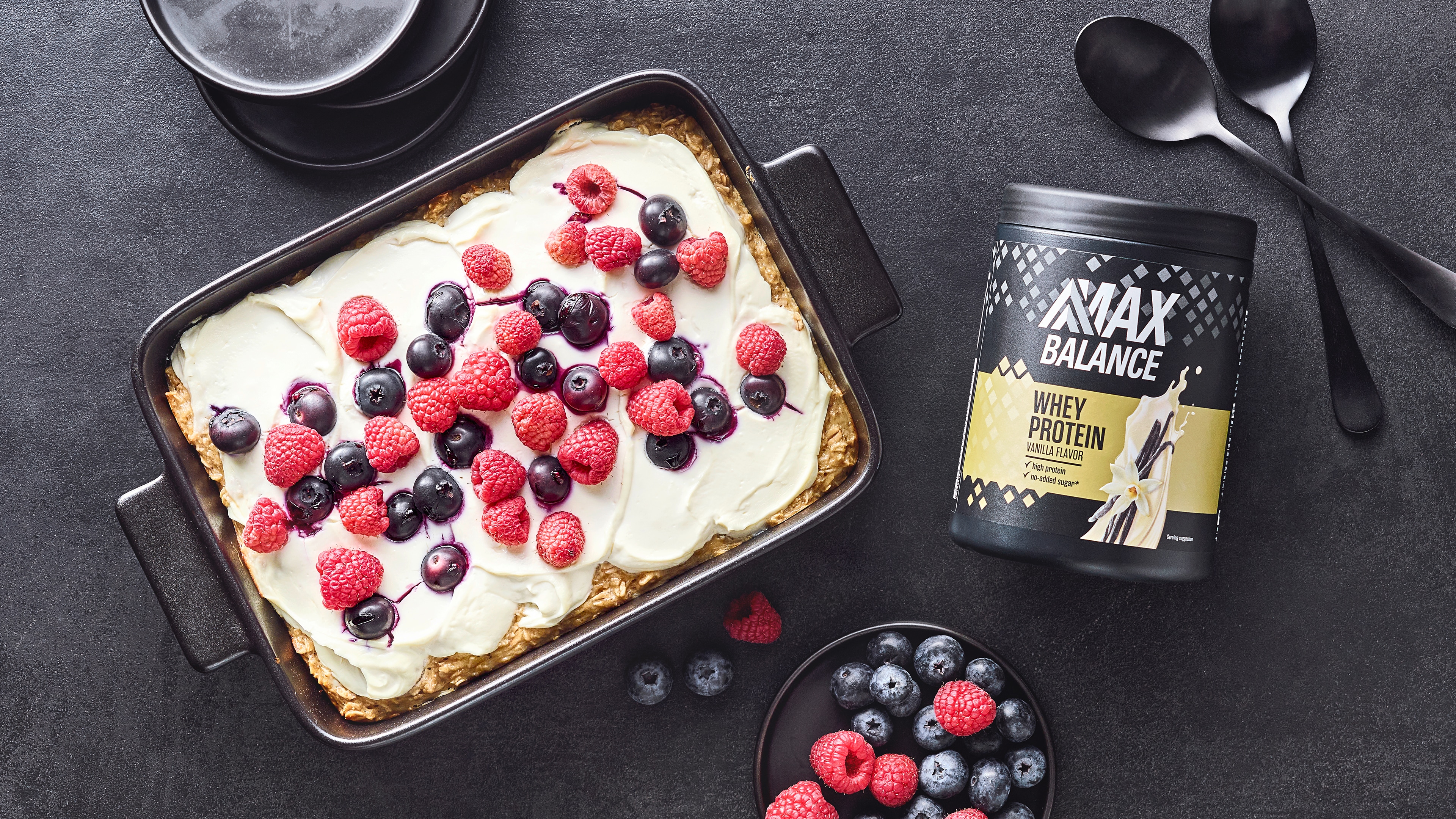 Auflaufform mit gebackenem Teig, heller Creme und Beeren, daneben eine Dose MaxBalance Proteinpulver Vanille