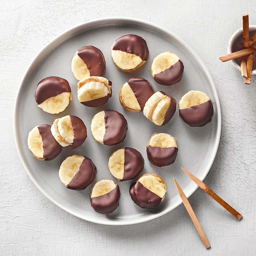 Kleine Banana Bites aus zwei Bananenscheiben, gefüllt mit Erdnussbutter und zur Hälfte in Schokolade getaucht.