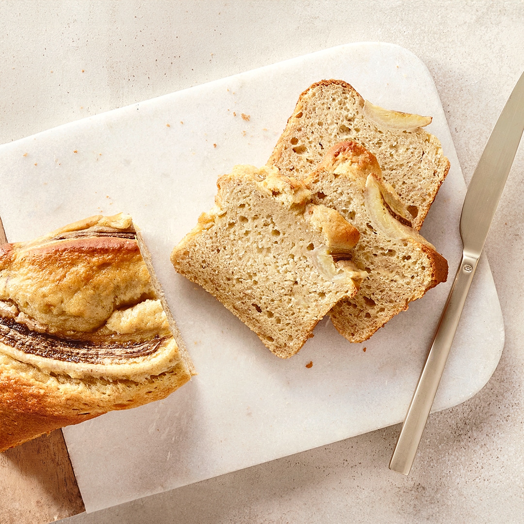 Ein Brett mit einem hellen Laib und drei Scheiben Bananenbrot, darin helle Stücke und eine halbe Banane auf der Oberfläche