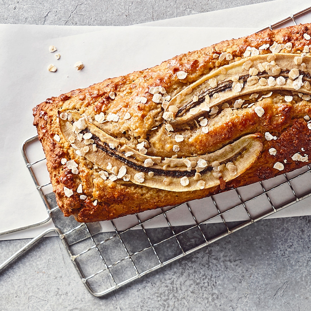 Ein Gitter mit einem goldbraunen Bananenbrot, garniert mit längs geschnittenen Bananenscheiben und groben Haferflocken