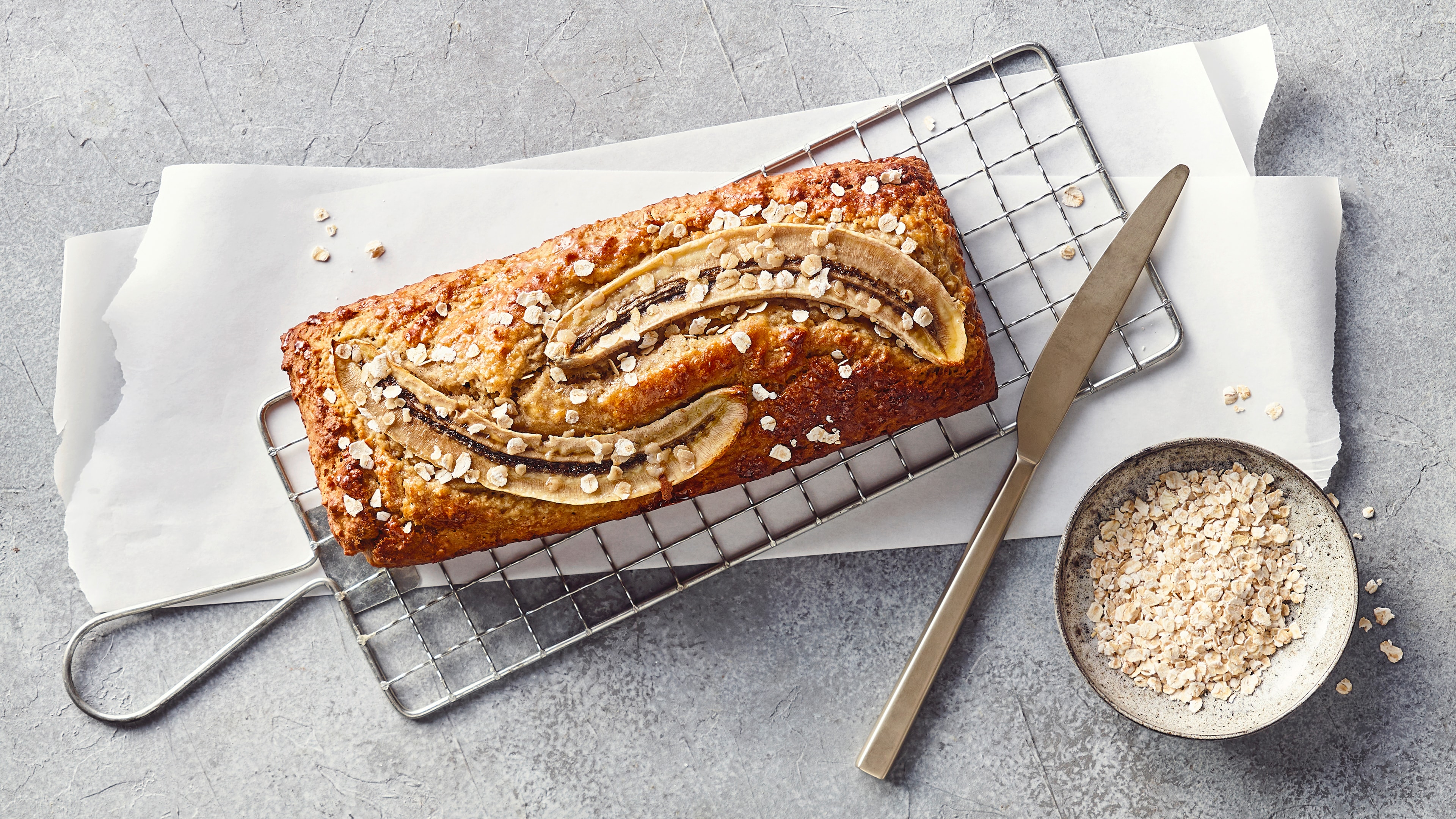 Ein Gitter mit einem goldbraunen Bananenbrot, garniert mit längs geschnittenen Bananenscheiben und groben Haferflocken