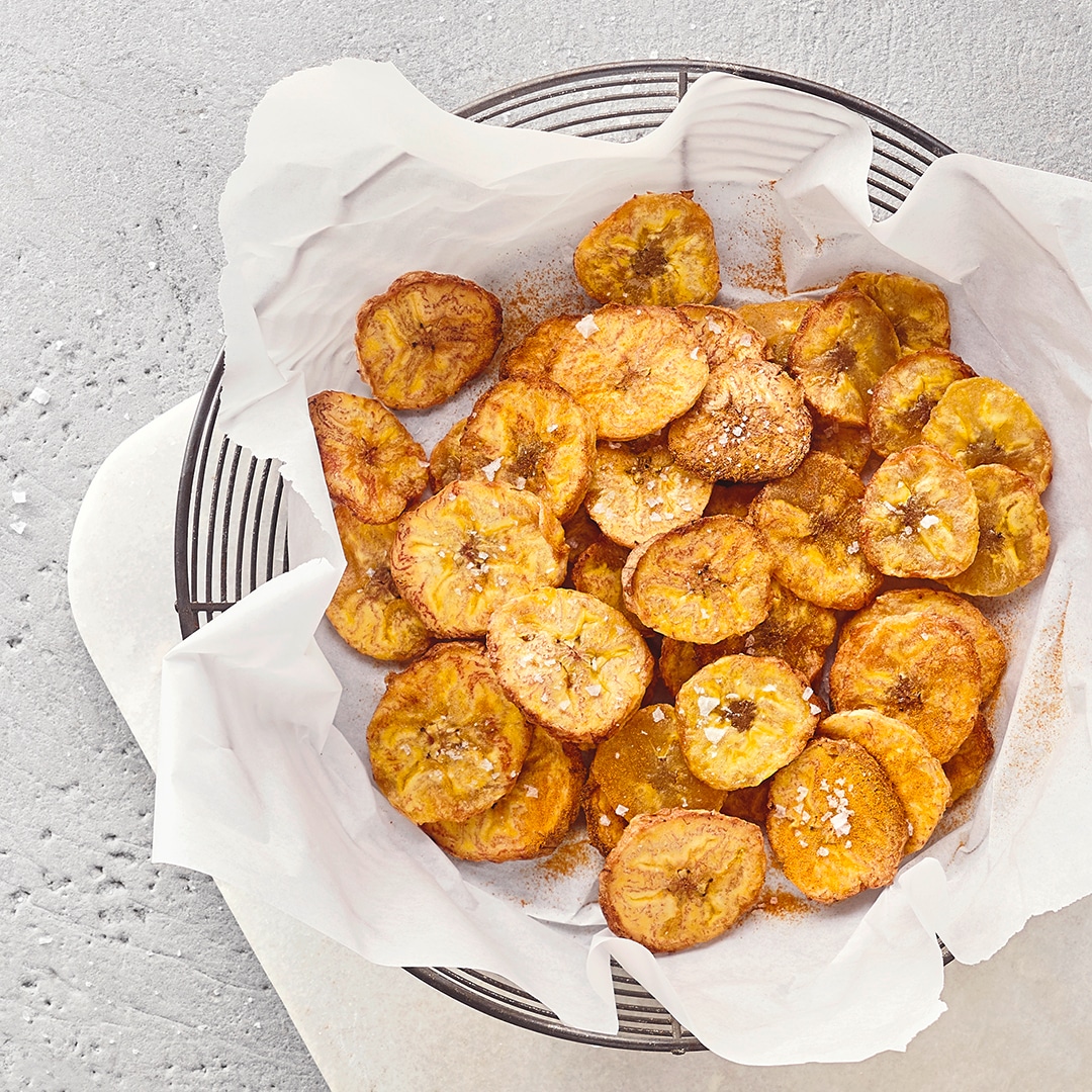 Metallschale aus Gittergeflecht, gefüllt mit knusprigen Bananenchips auf einer Serviette, auf einem Brett platziert.