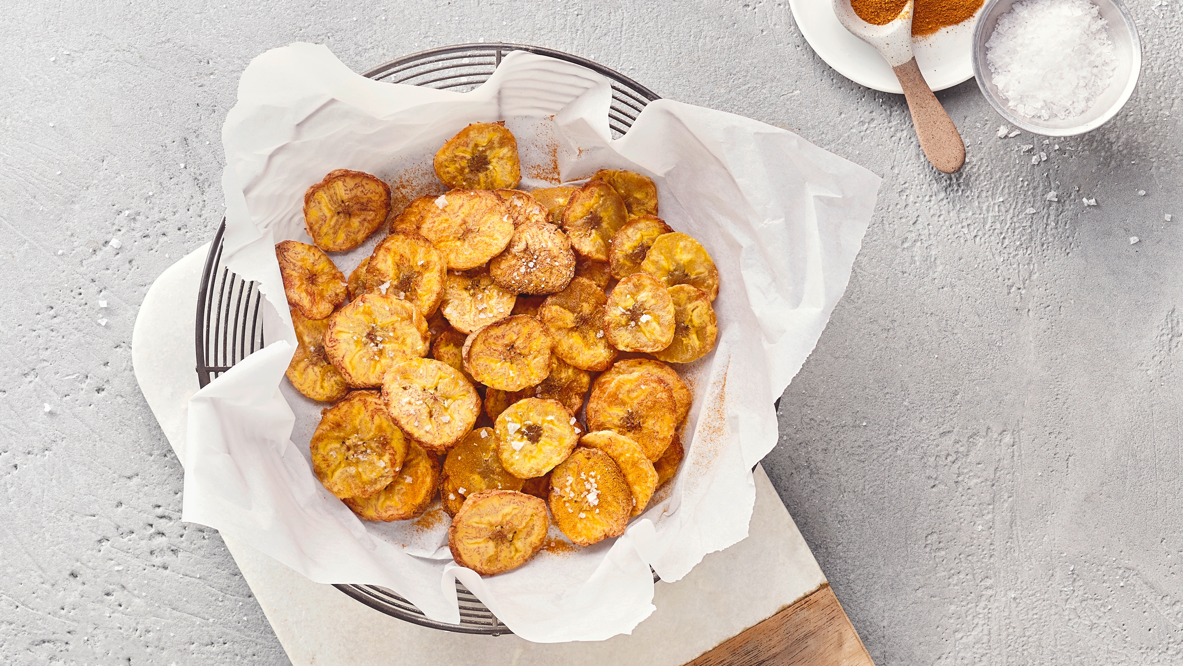 Metallschale aus Gittergeflecht, gefüllt mit knusprigen Bananenchips auf einer Serviette, auf einem Brett platziert. Rechts oben steht etwas Zimt und Zucker.