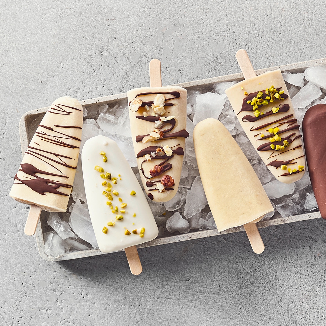 Ein Tablett mit hellen Eis am Stiel, teils mit dunkler Schokolade, Nüssen oder grünen Pistazienstücken verziert, auf Eiswürfeln.