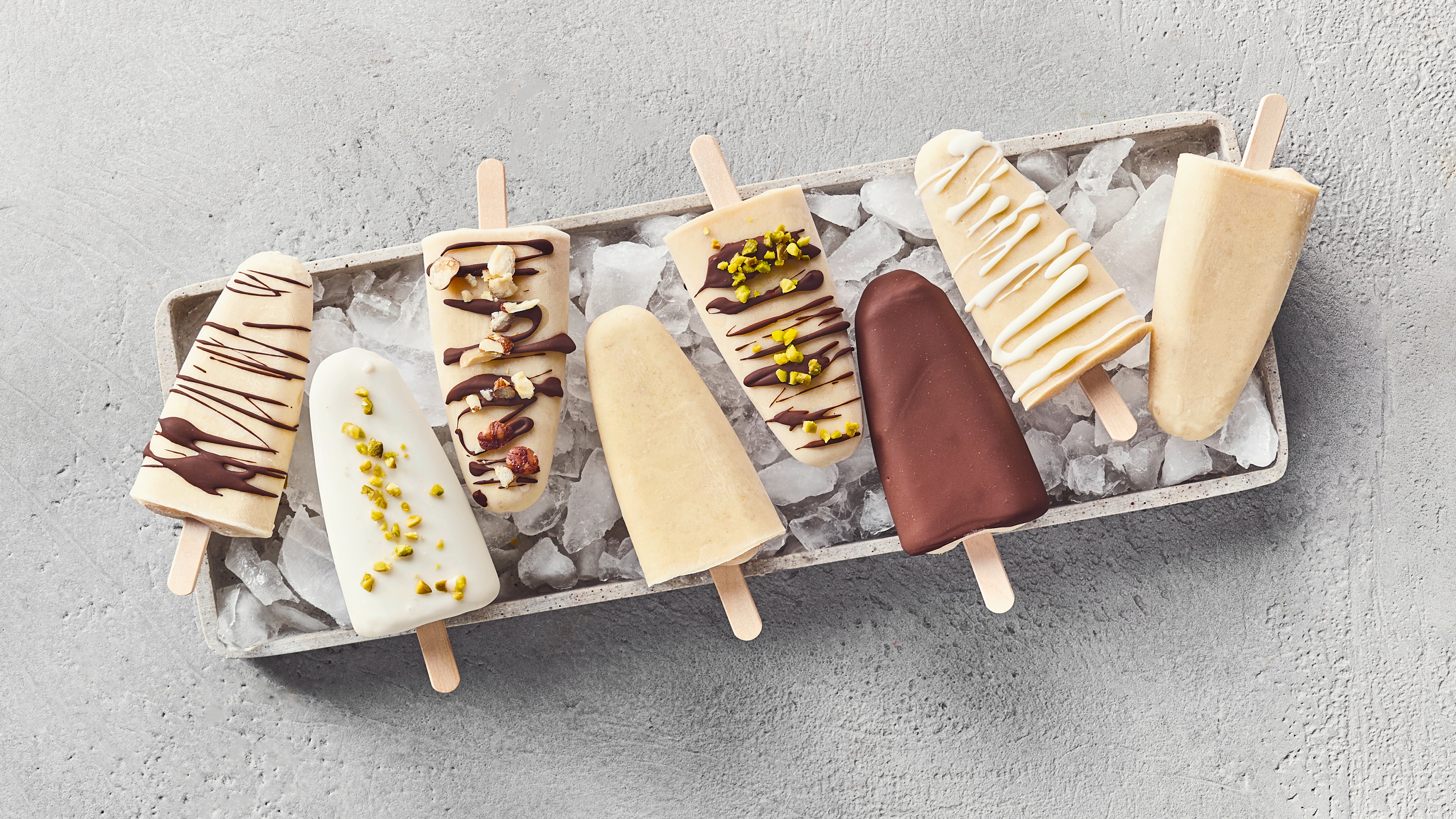 Ein Tablett mit hellen Eis am Stiel, teils mit dunkler Schokolade, Nüssen oder grünen Pistazienstücken verziert, auf Eiswürfeln.