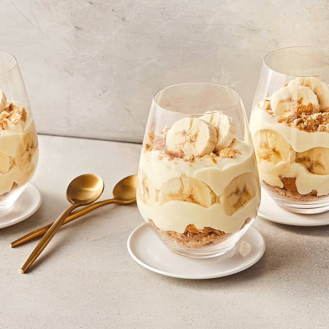Bauchige Dessertgläser, befüllt mit Schichten aus knusprigen Butterkeksstückchen, heller Creme und Bananenscheiben