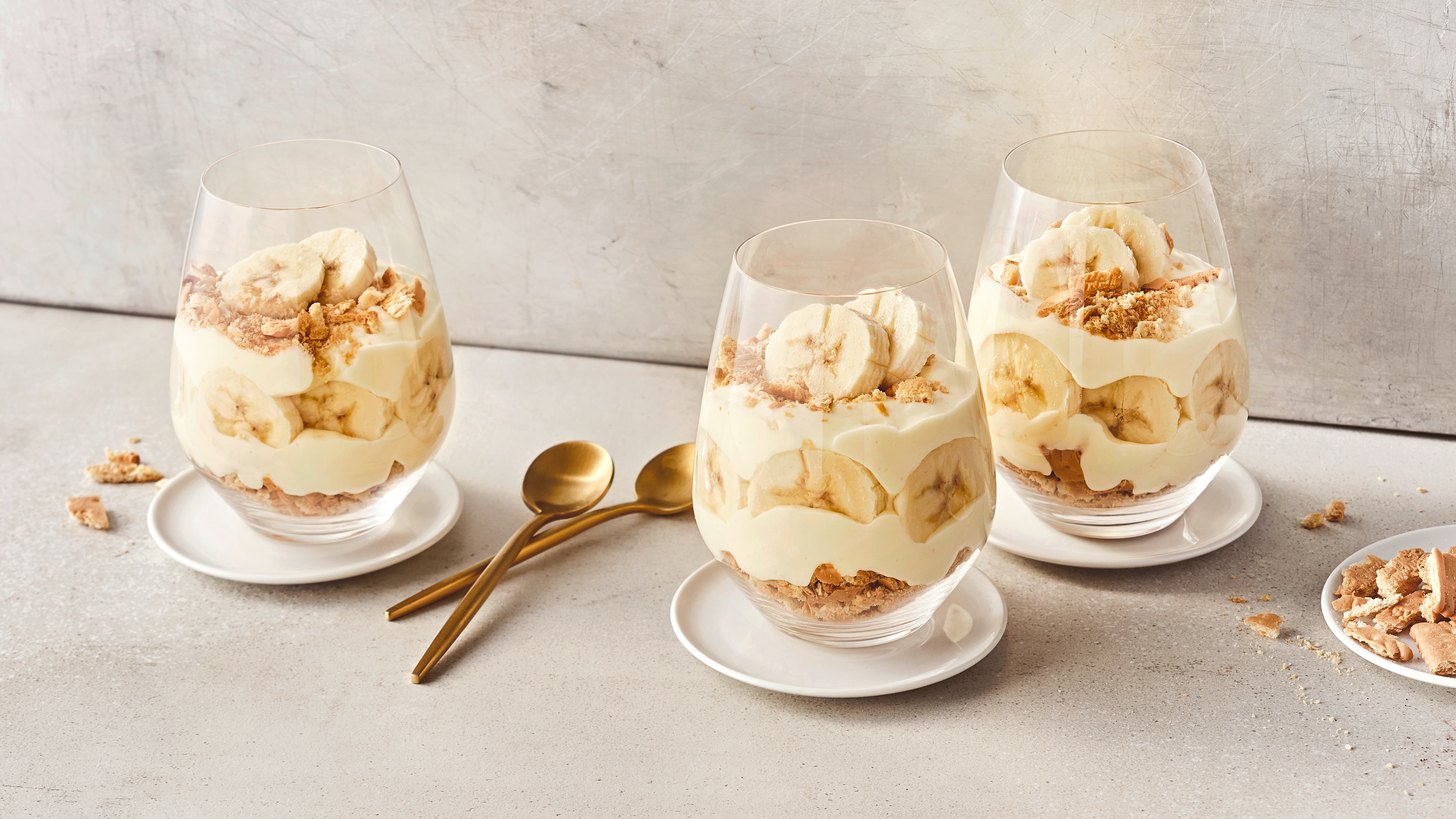 Dessert-Rezept für Bananenpudding Bauchige Dessertgläser, befüllt mit Schichten aus knusprigen Butterkeksstückchen, heller Creme und Bananenscheiben