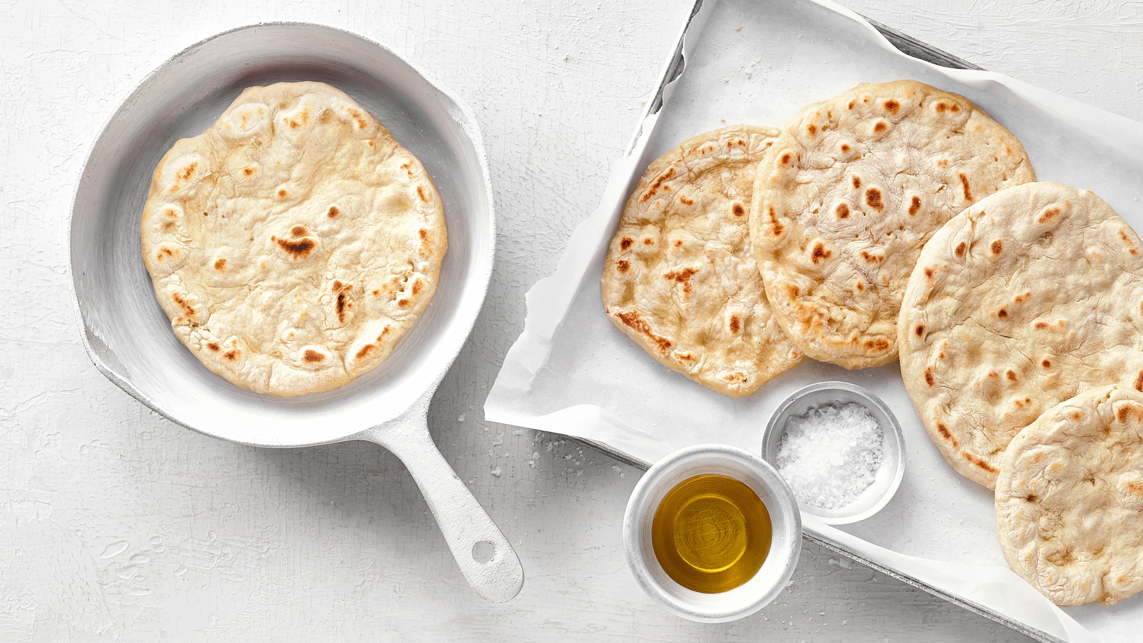 Pfanne mit einem kleinen, runden Fladenbrot. Rechts daneben ein Backblech mit vier weiteren Fladenbroten, einer kleinen Salzschale und einer Ölschale.