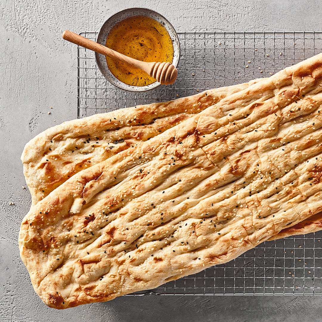 Ein Gitter mit einem langen, hellen Fladenbrot, das leicht gebräunt ist und schwarze sowie weiße Samen trägt, daneben eine Schale mit gelber Flüssigkeit und einem Holzstab.