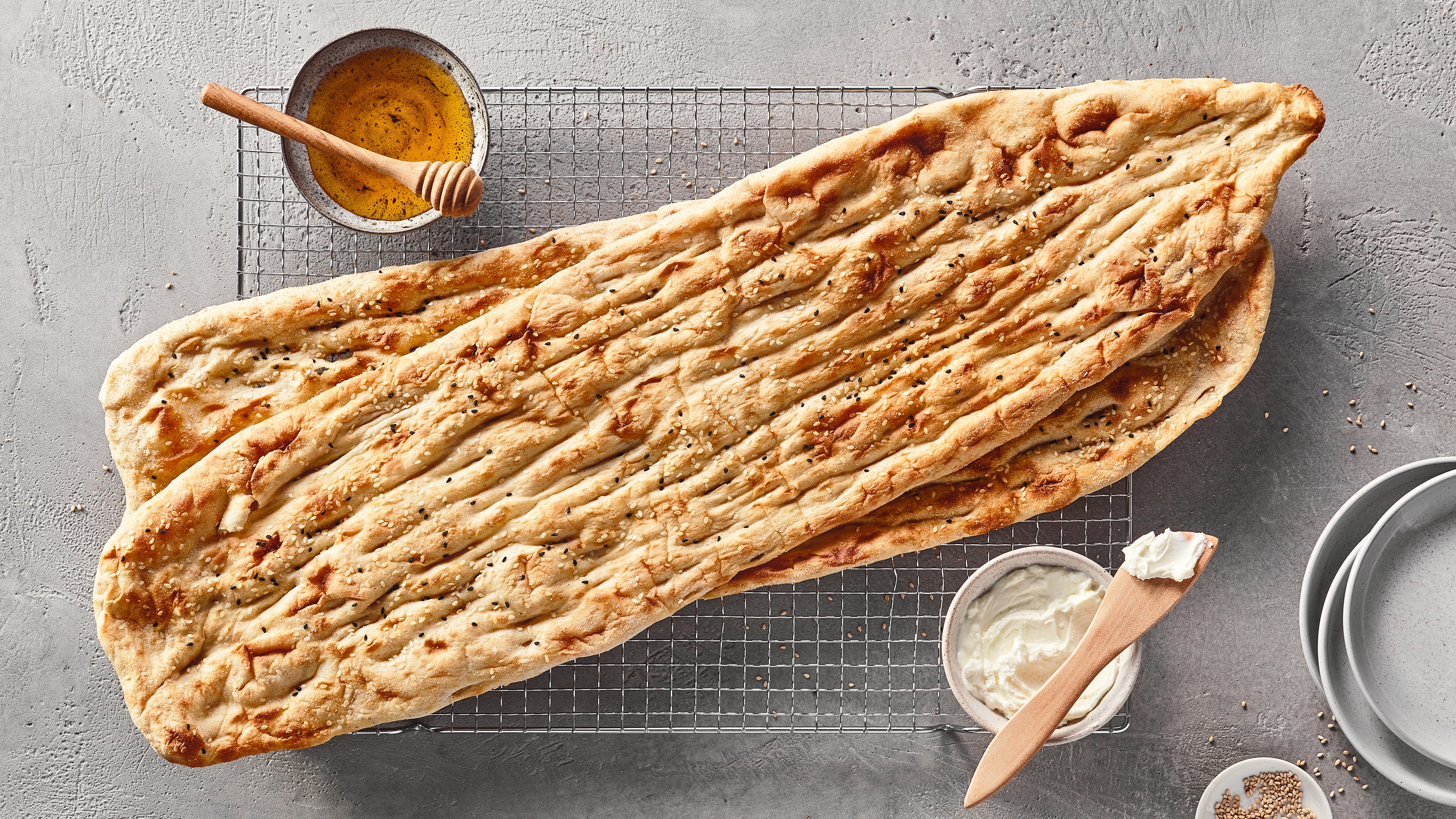 Ein Gitter mit einem langen, hellen Fladenbrot, das leicht gebräunt ist und schwarze sowie weiße Samen trägt, daneben eine Schale mit gelber Flüssigkeit und einem Holzstab.
