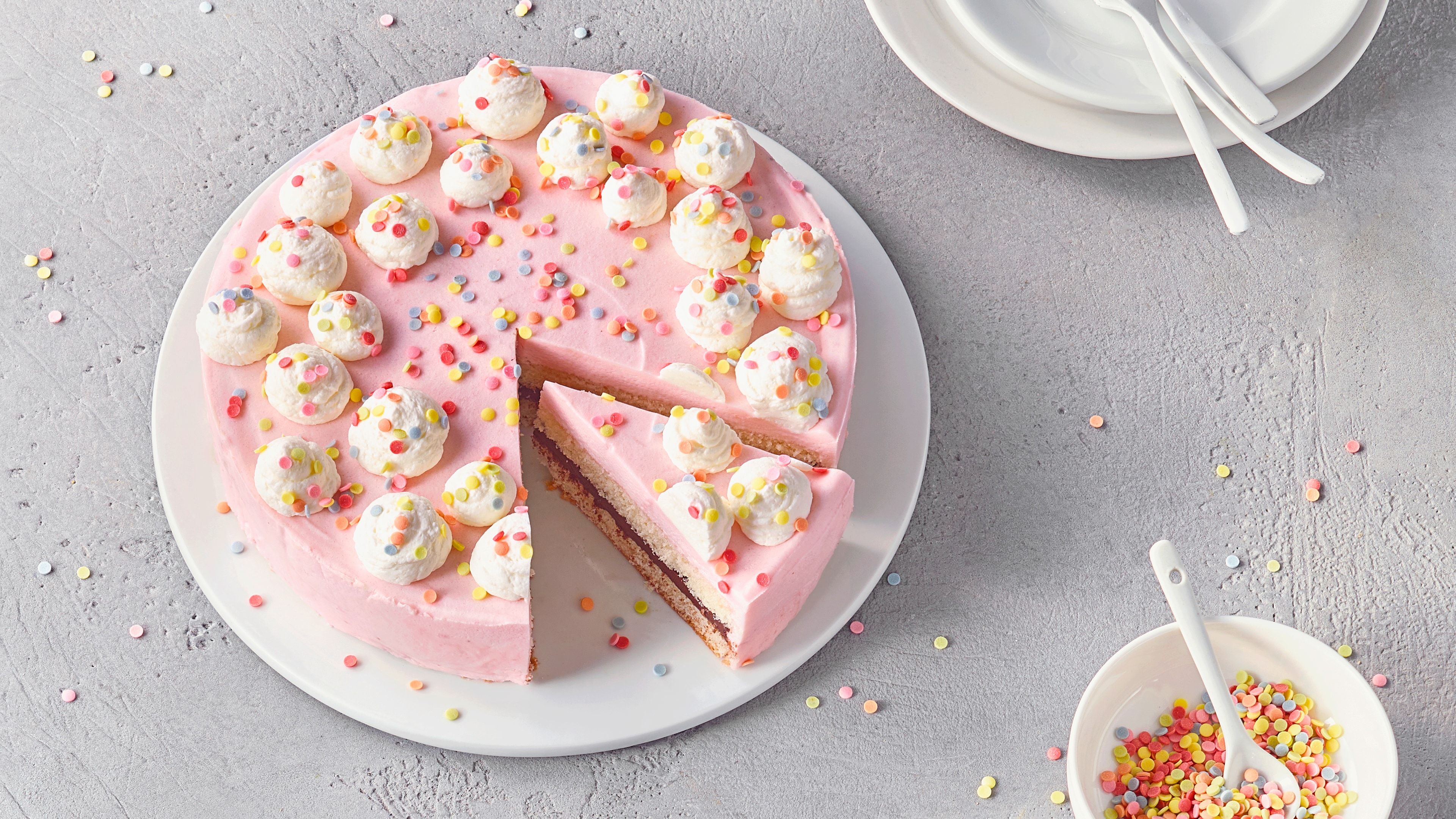 Ein Teller mit einer rosa Cremetorte, garniert mit weißen Cremetupfen und bunten Zuckerstreuseln, ein Stück herausgeschnitten.