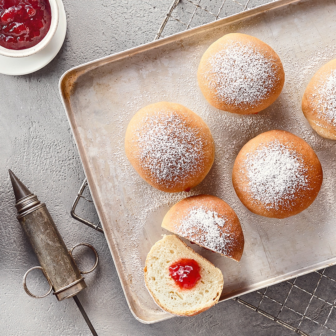 Ein Backblech mit gebackenen Teigstückchen, davon eins aufgeschnitten & mit roter Marmelade gefüllt, garniert mit Puderzucker