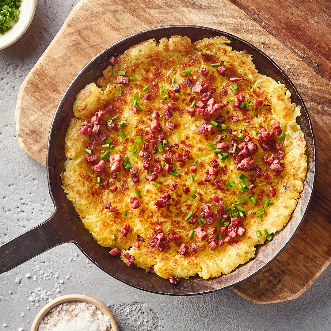 Eine Pfanne mit einem großen goldbraunen Rösti, belegt mit vielen kleinen Speckwürfeln und grünen Schnittlauchringen