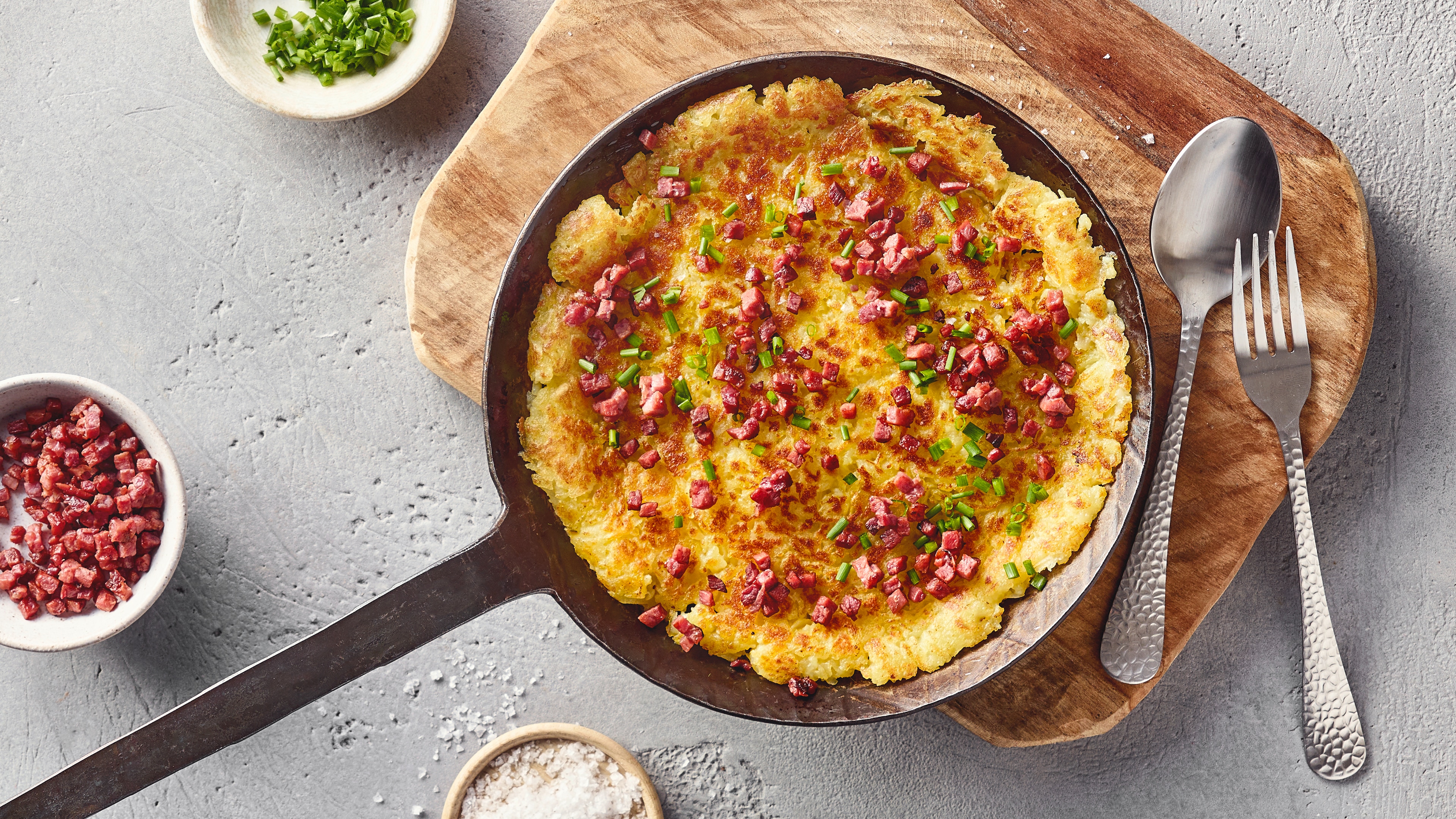 Eine Pfanne mit einem großen goldbraunen Rösti, belegt mit vielen kleinen Speckwürfeln und grünen Schnittlauchringen