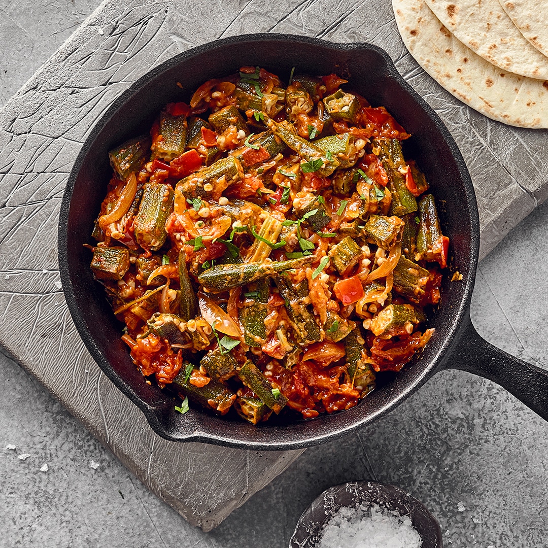 Eine Pfanne mit Okra, Tomaten und Zwiebeln, daneben Tortillas, gehackte Kräuter und eine Schale mit grobem Salz