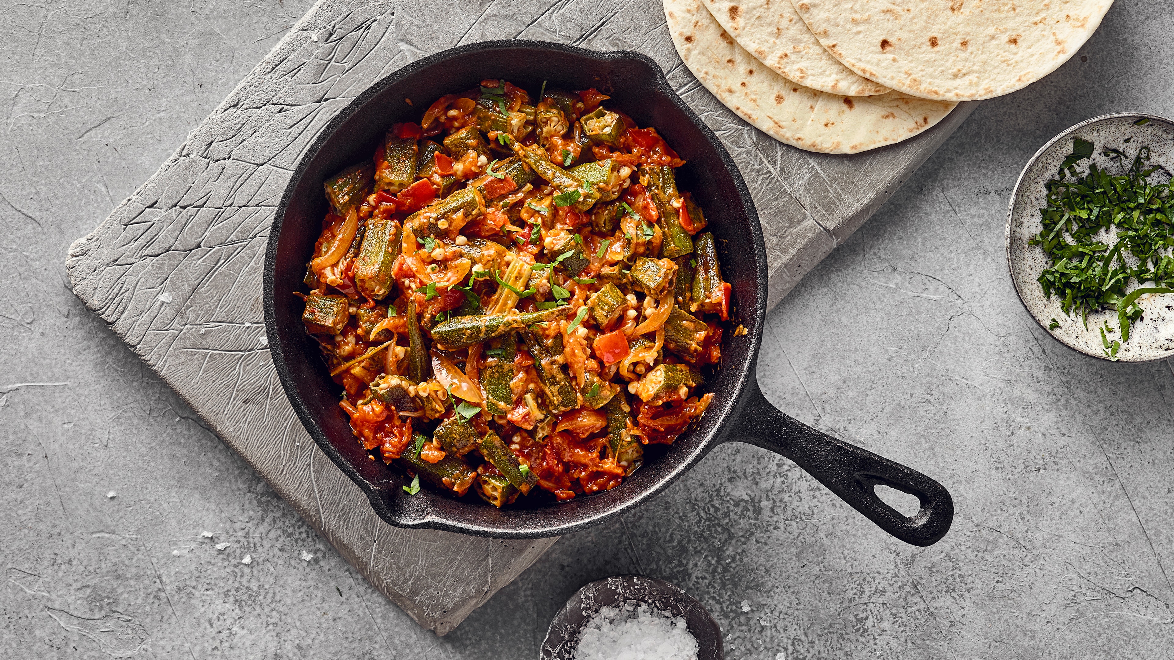 Rezept für Bhindi Masala Eine Pfanne mit Okra, Tomaten und Zwiebeln, daneben Tortillas, gehackte Kräuter und eine Schale mit grobem Salz