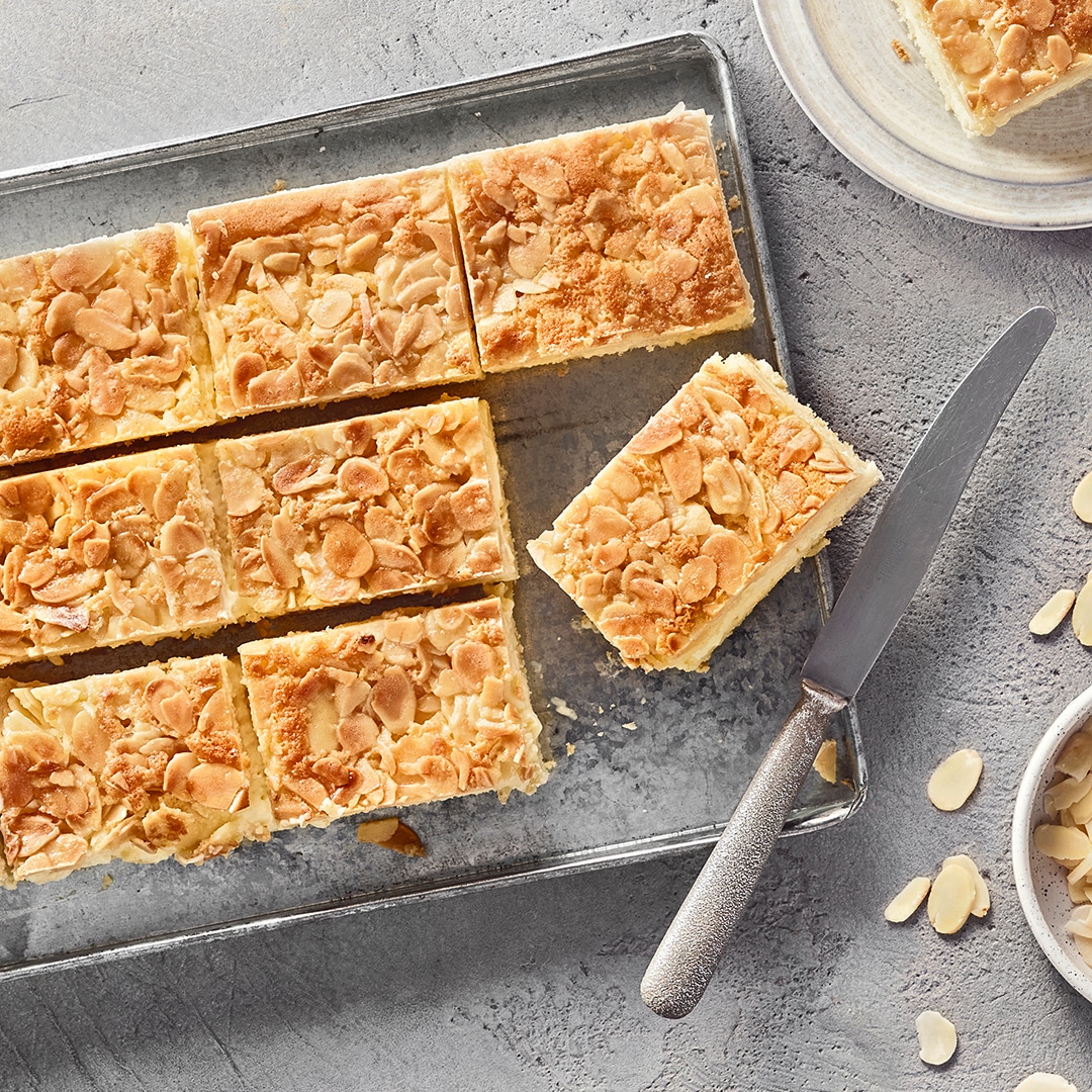 Ein Blech mit rechteckigen Stücken, hellen Schichten und einer Oberfläche aus dünnen, hellbraunen Scheiben und Krümeln.