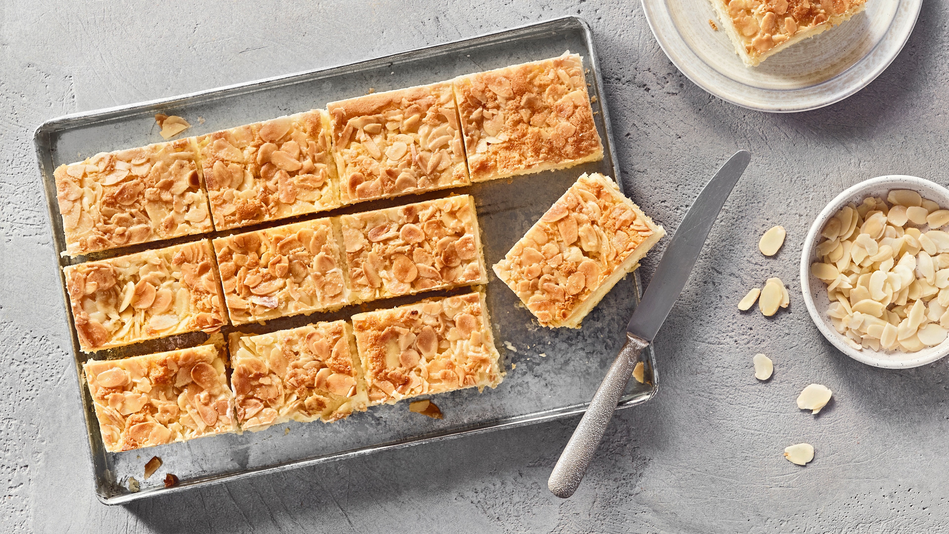 Ein Blech mit rechteckigen Stücken, hellen Schichten und einer Oberfläche aus dünnen, hellbraunen Scheiben und Krümeln.