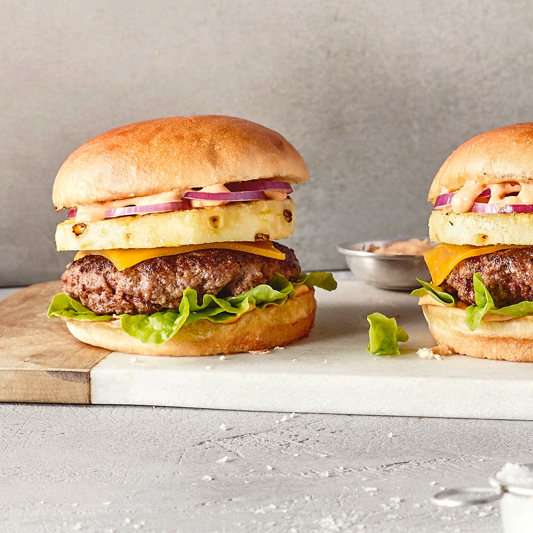 Ein Brett mit zwei Burgern, belegt mit Salat, braunem Patty, gelbem Käse, Ananasscheibe und roten Zwiebelringen.