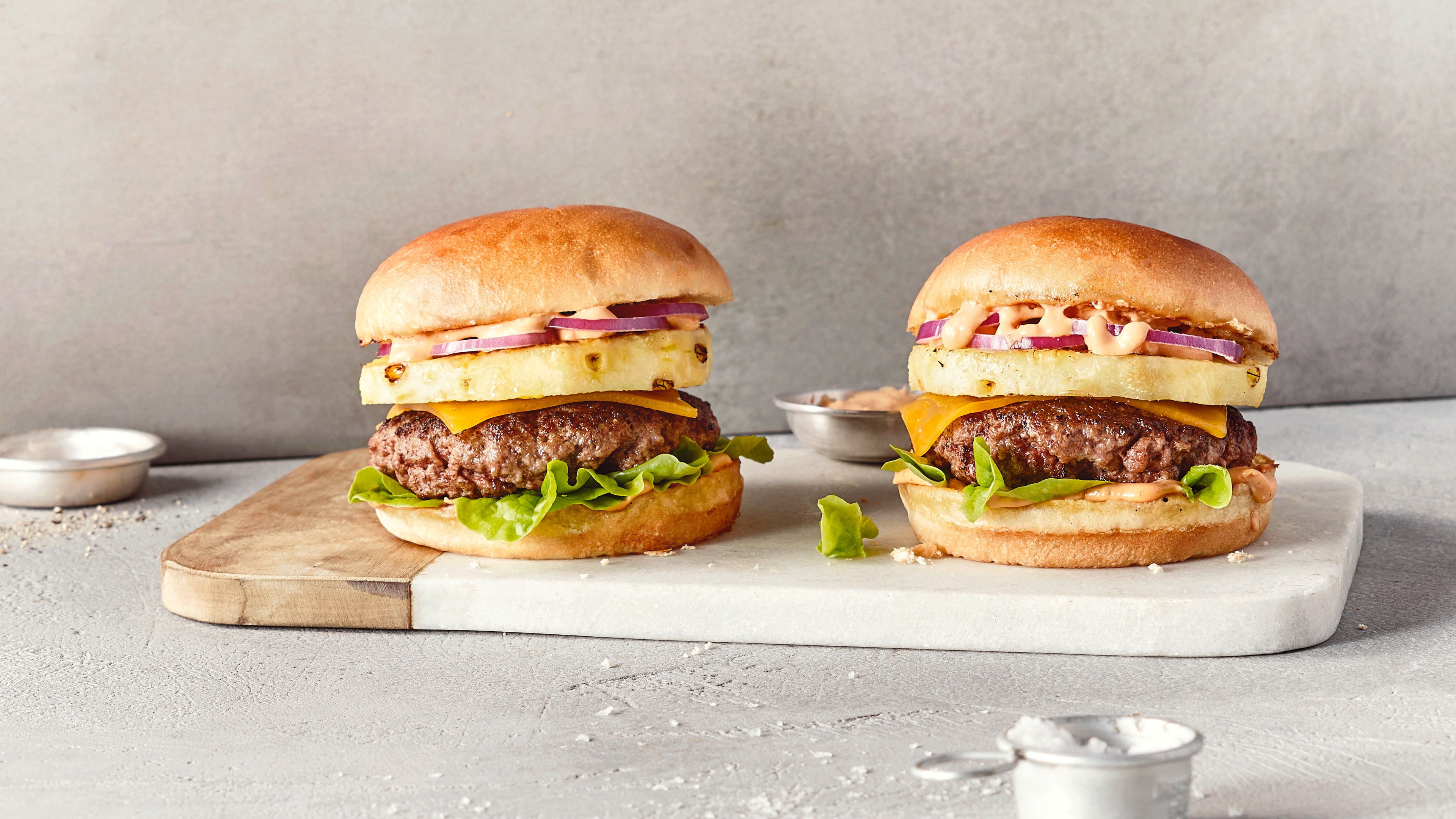 Ein Brett mit zwei Burgern, belegt mit Salat, braunem Patty, gelbem Käse, Ananasscheibe und roten Zwiebelringen.