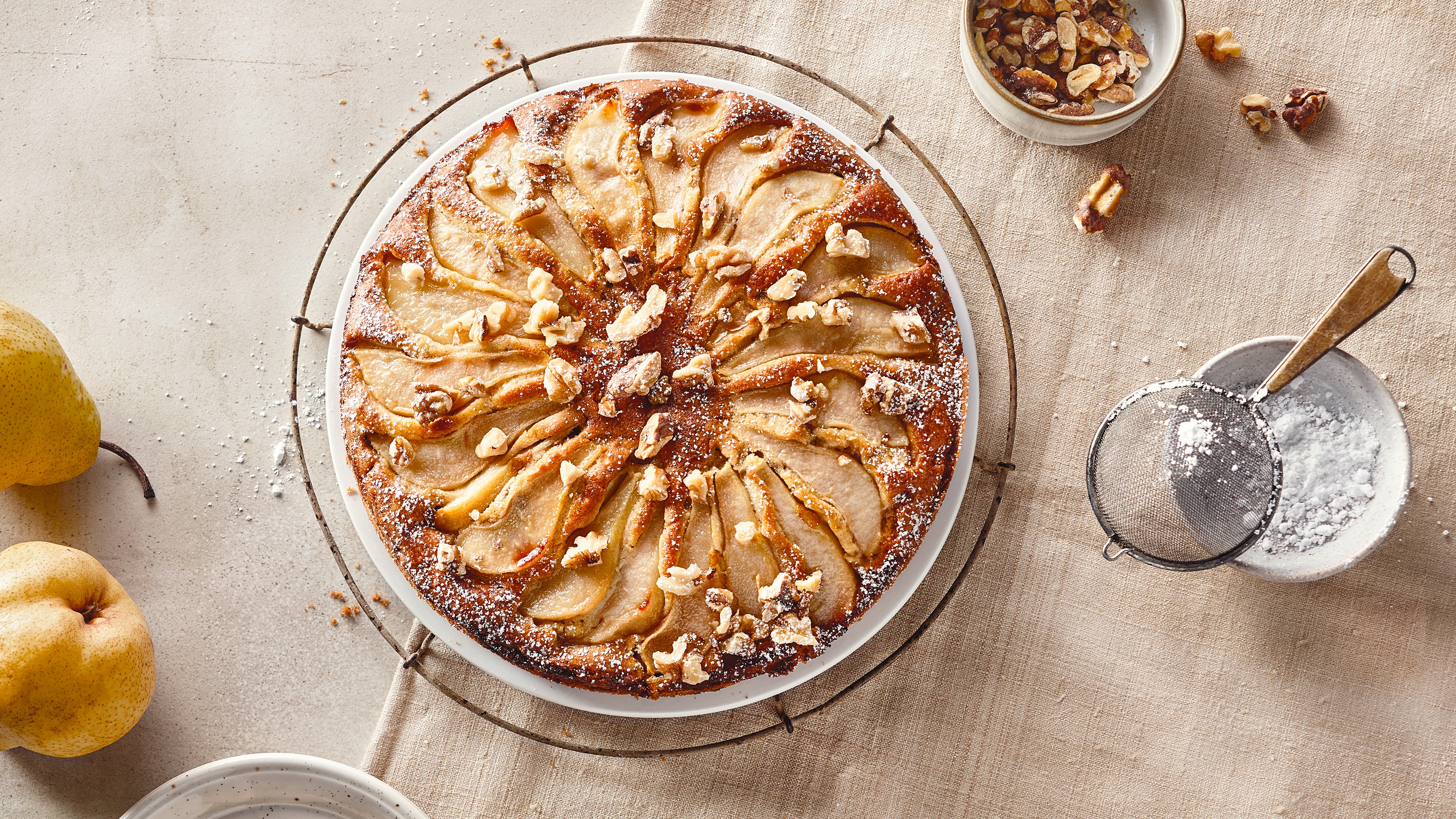 Rezept für Birnen-Walnuss-Kuchen Goldgelb gebackener Kuchen, getoppt mit Birnen und gehackten Walnüssen, bestreut mit Puderzucker, auf einem Abkühlgitter