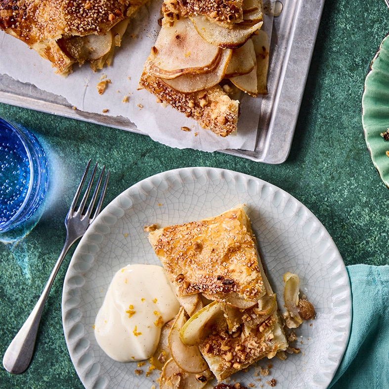 Stücke eines Birnenstrudels mit heller Quarkcreme auf Teller und Servierplatte.