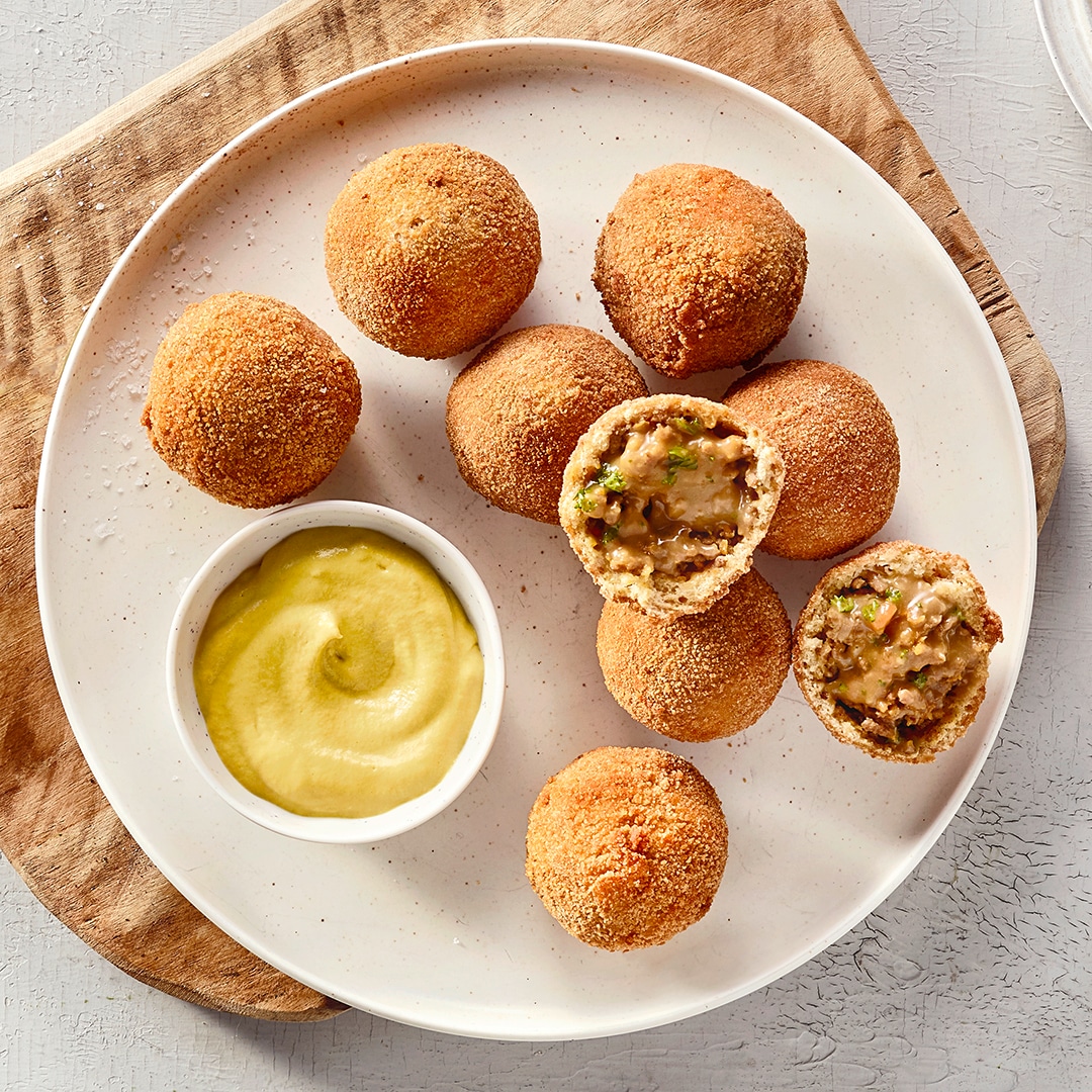 Ein Teller mit panierten Bällchen, zwei halb geöffnet mit heller Füllung und grünen Stücken, daneben gelbe Creme in einer Schale.