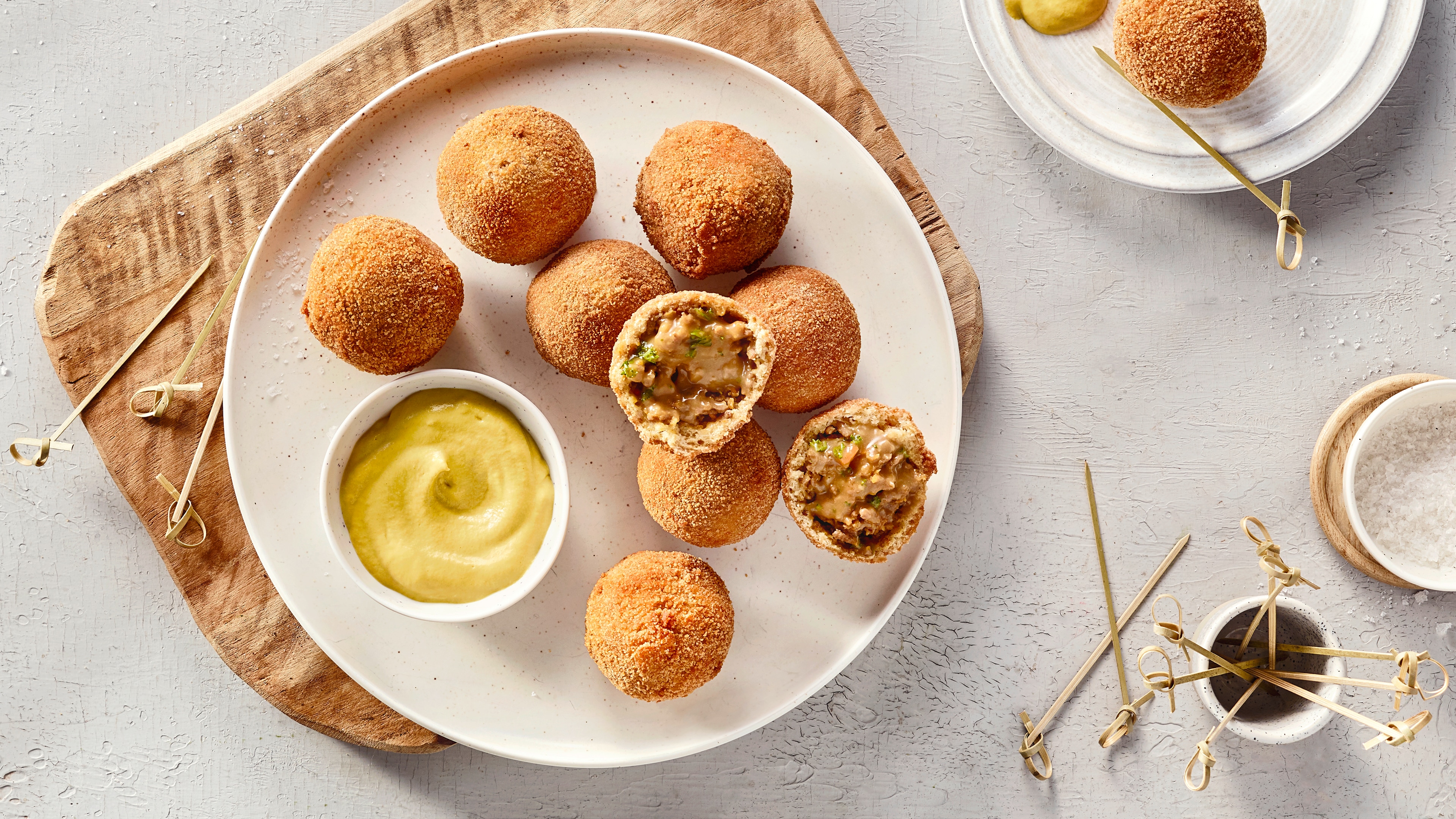 Ein Teller mit panierten Bällchen, zwei halb geöffnet mit heller Füllung und grünen Stücken, daneben gelbe Creme in einer Schale.