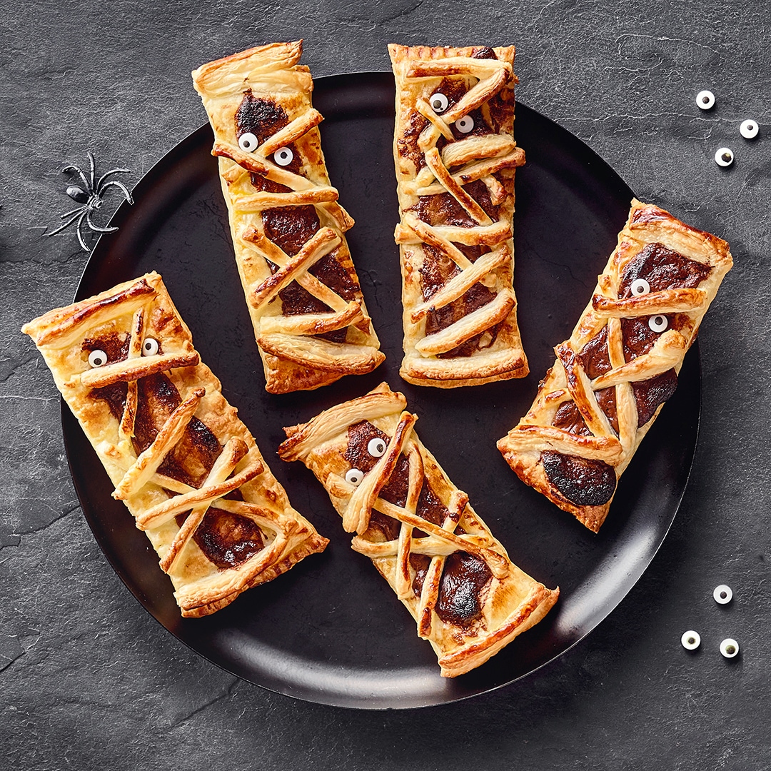 Blätterteig-Snacks als Mumien dekoriert mit Zuckerguss-Augen auf Tellern, Halloween-Stil mit kleinen Kunststoffspinnen.