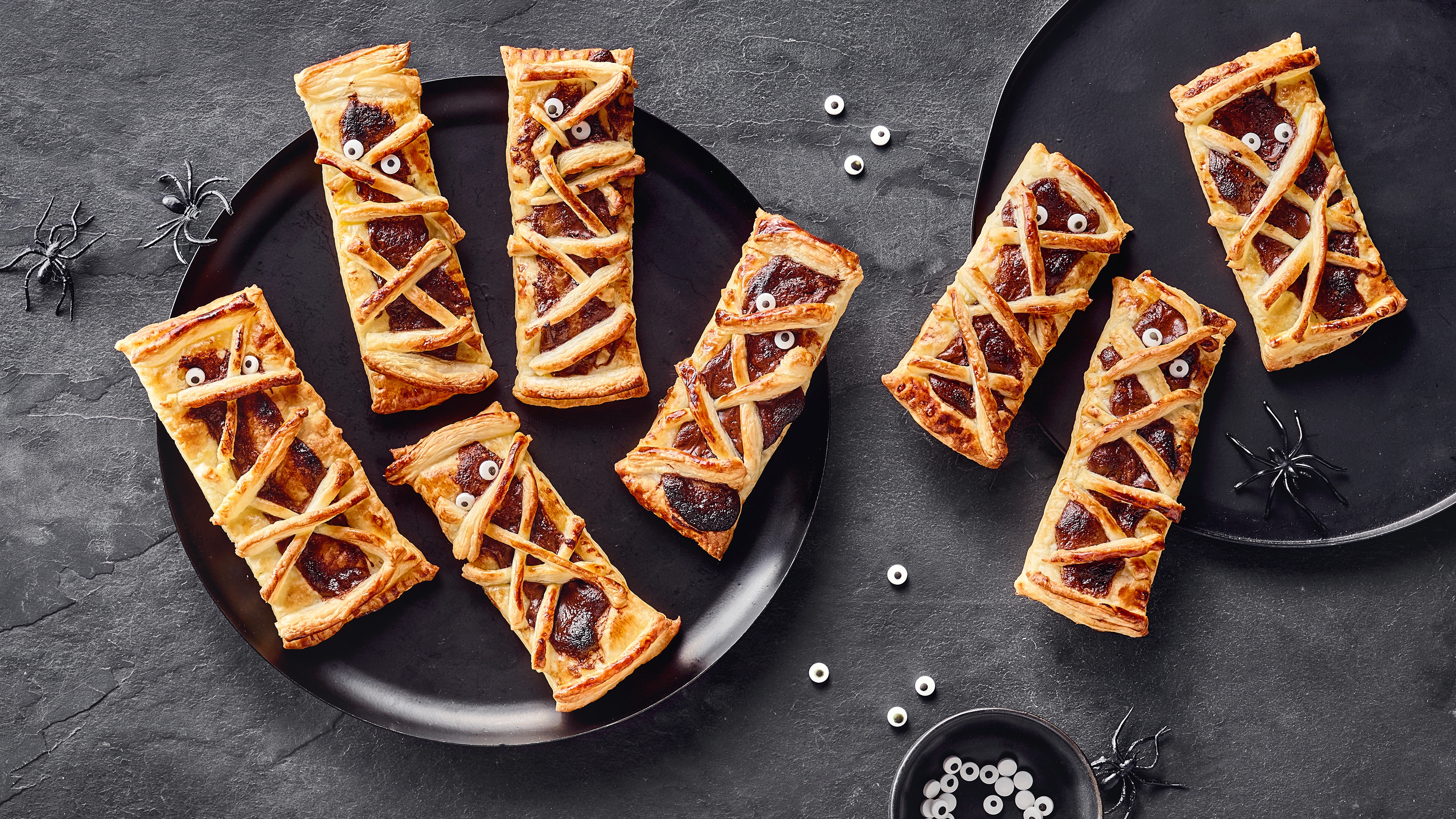 Blätterteig-Snacks als Mumien dekoriert mit Zuckerguss-Augen auf Tellern, Halloween-Stil mit kleinen Kunststoffspinnen.