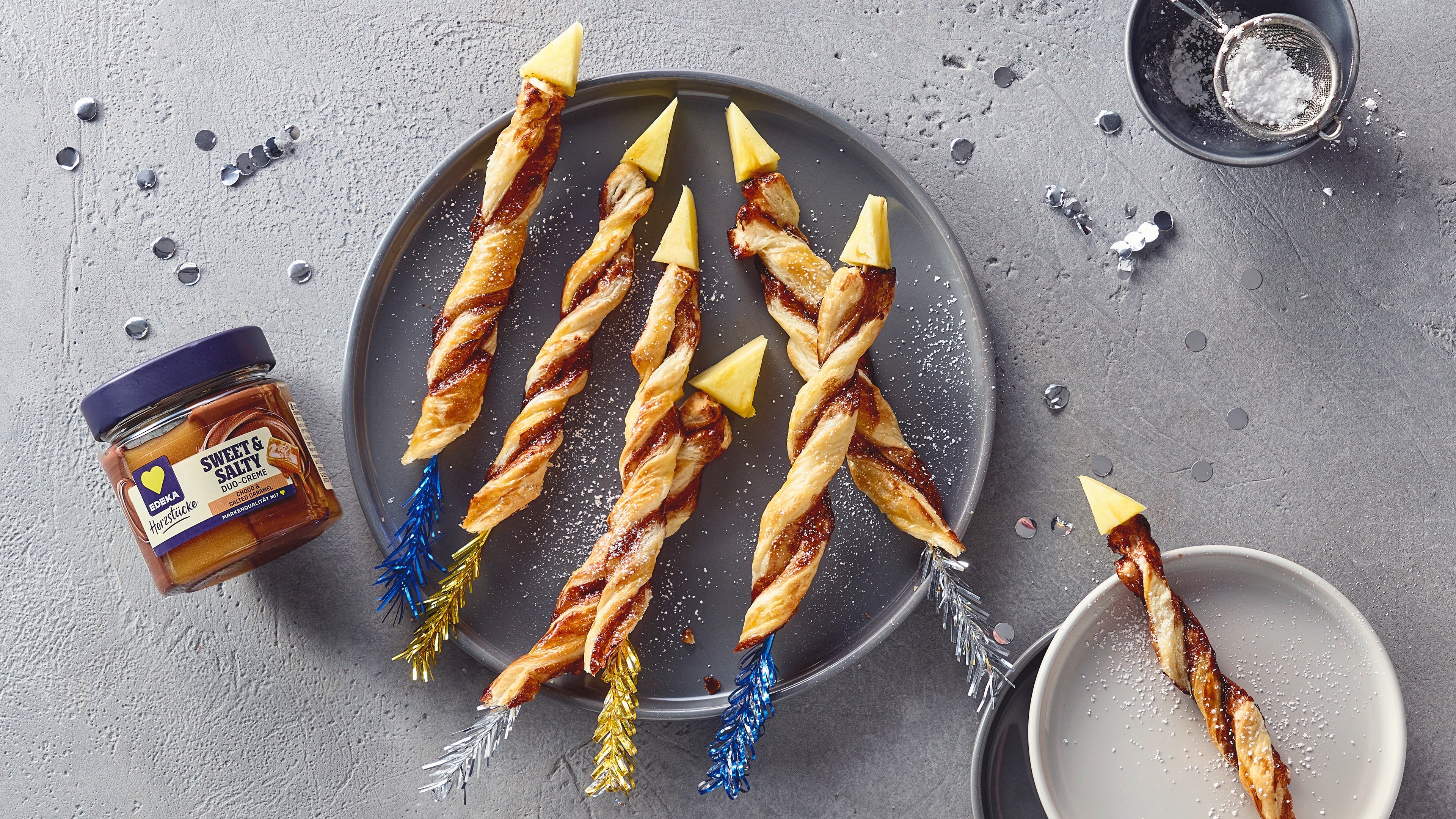 Ein Teller mit sechs gedrehten Blätterteigstangen, gefüllt mit dunkler Creme, bestreut mit Zucker und gelben Spitzen