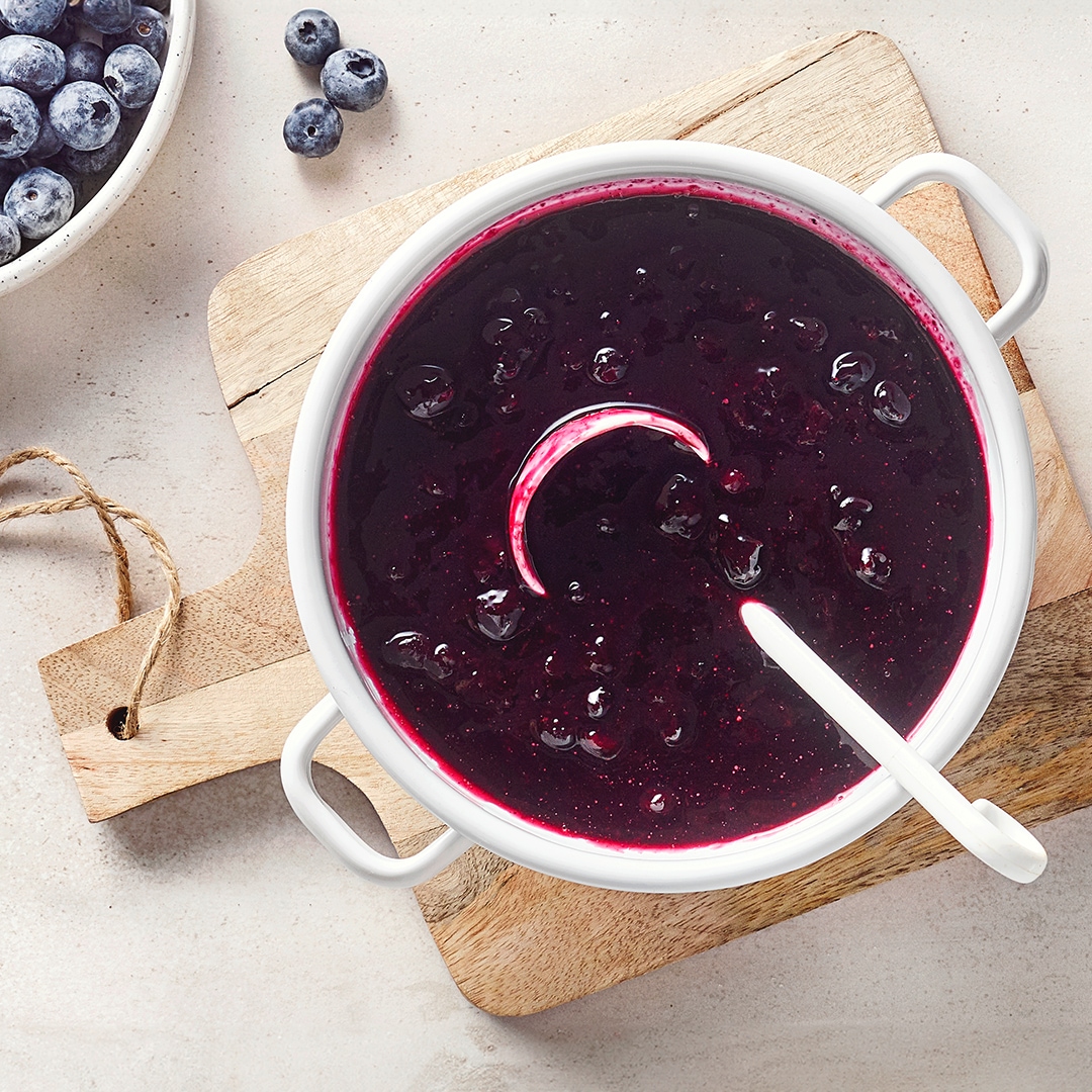 Ein Topf mit dunkler violetter Soße, darin runde Früchte, am Rand ein weißer Löffel und daneben einzelne blaue Beeren.