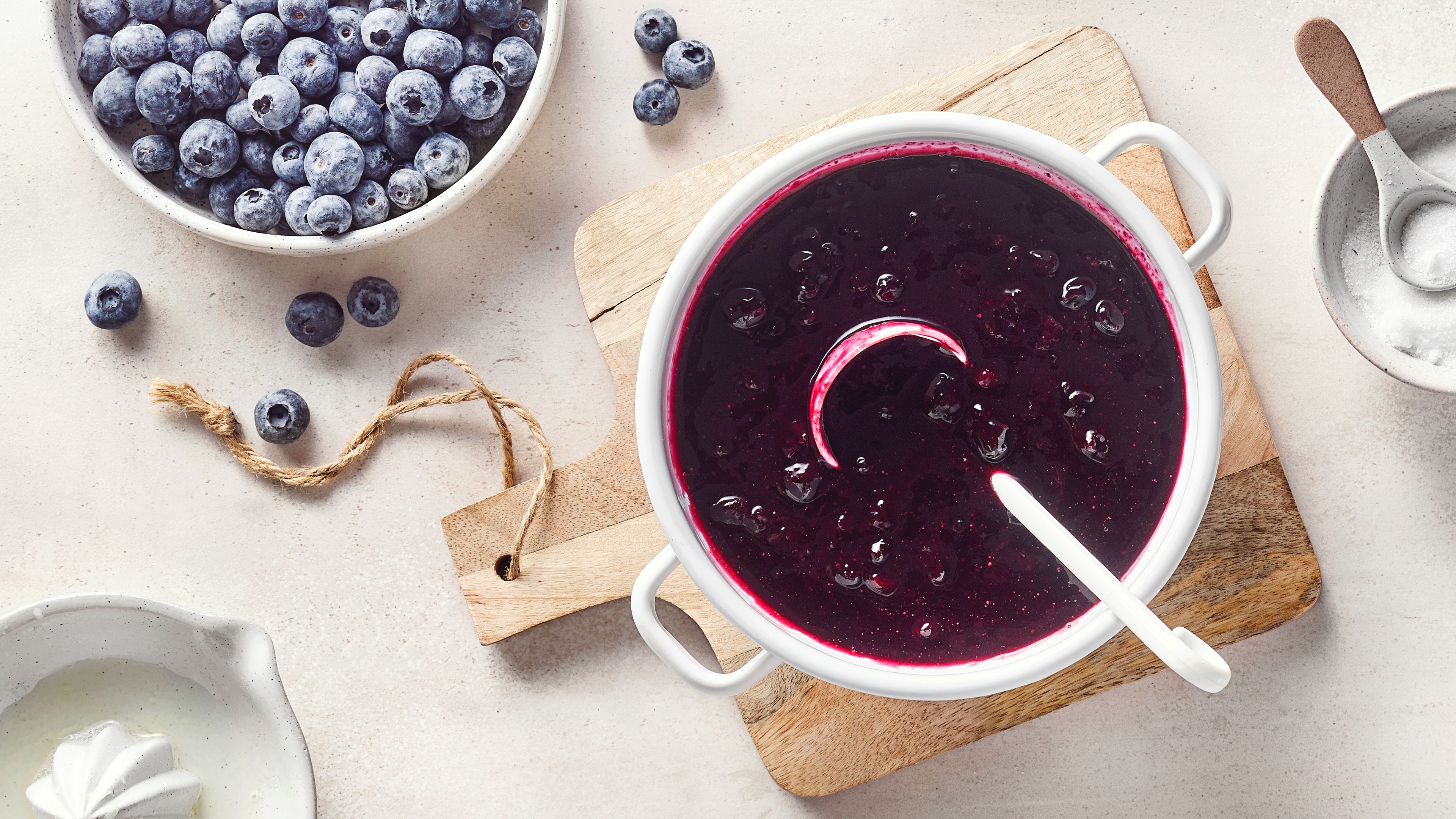 Ein Topf mit dunkler violetter Soße, darin runde Früchte, am Rand ein weißer Löffel und daneben einzelne blaue Beeren.