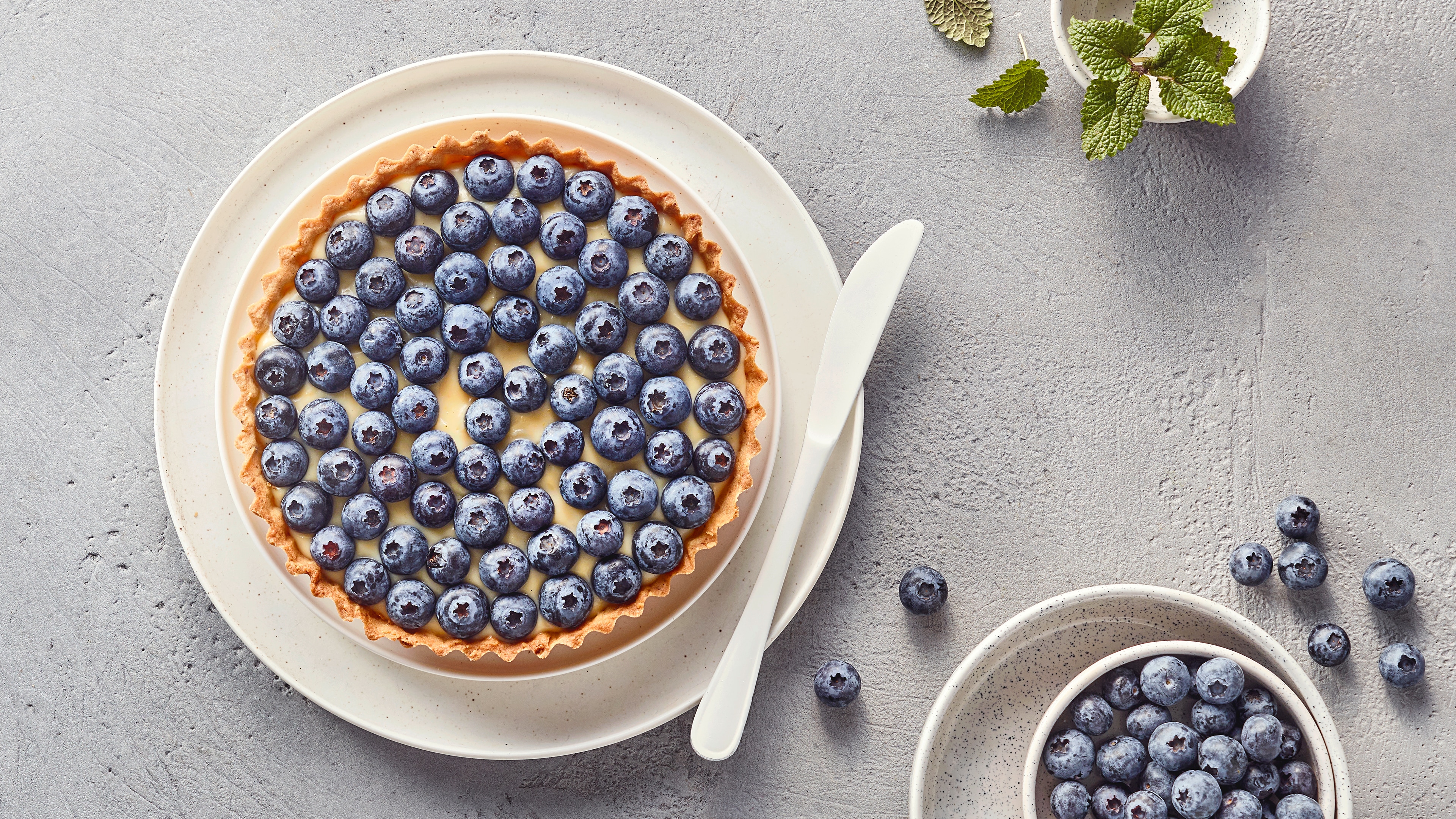 Runde Tarte mit cremiger Füllung und Blaubeeren, daneben Schälchen mit Blaubeeren
