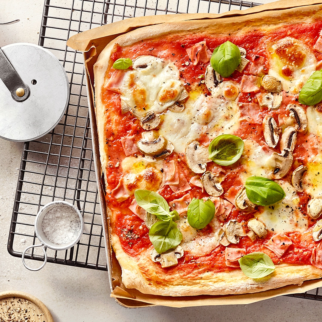 Ein Blech mit rechteckiger Pizza, belegt mit roter Soße, geschmolzenem Käse, Schinken, Pilzscheiben und grünen Basilikumblättern.