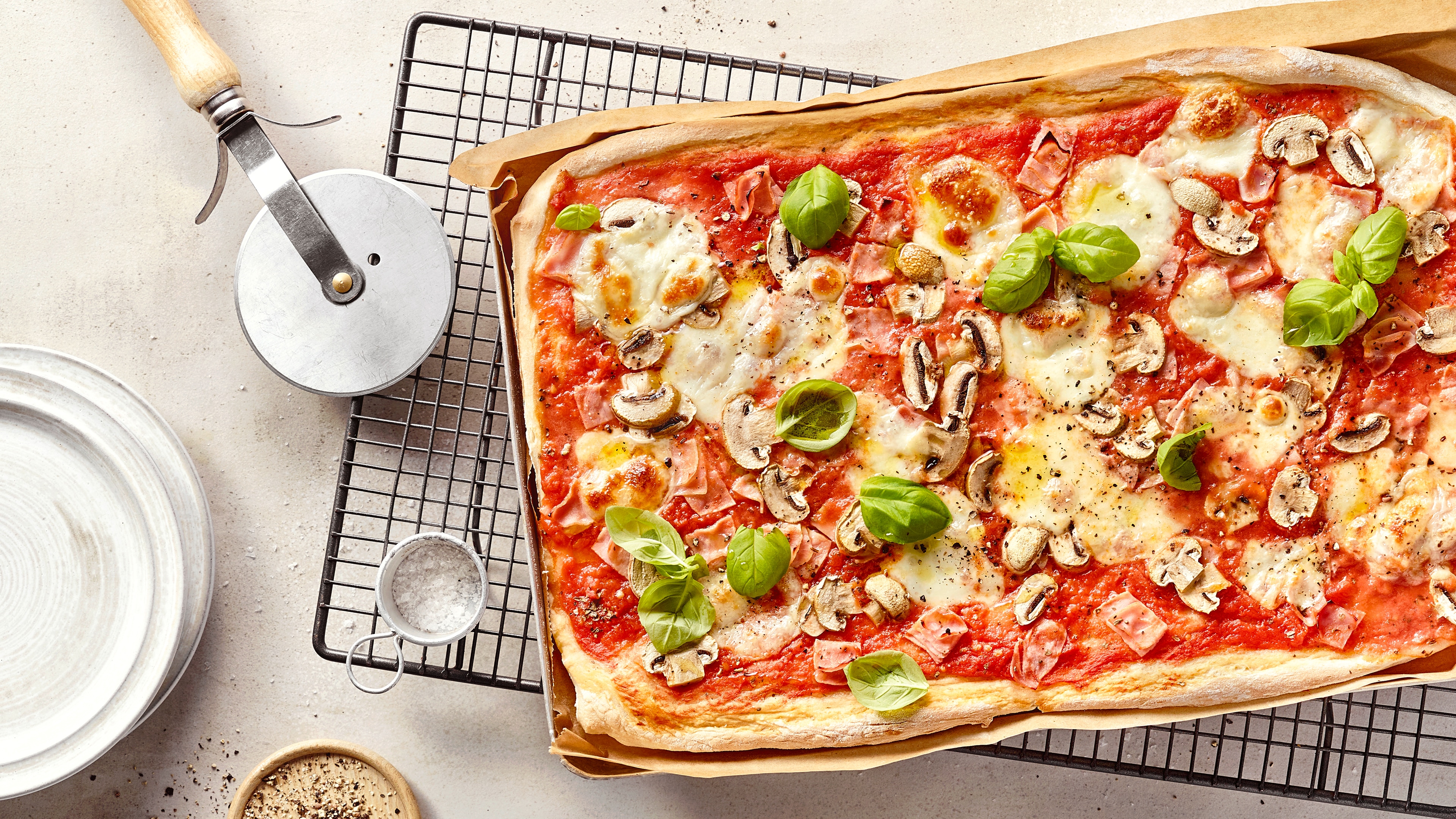 Ein Blech mit rechteckiger Pizza, belegt mit roter Soße, geschmolzenem Käse, Schinken, Pilzscheiben und grünen Basilikumblättern.