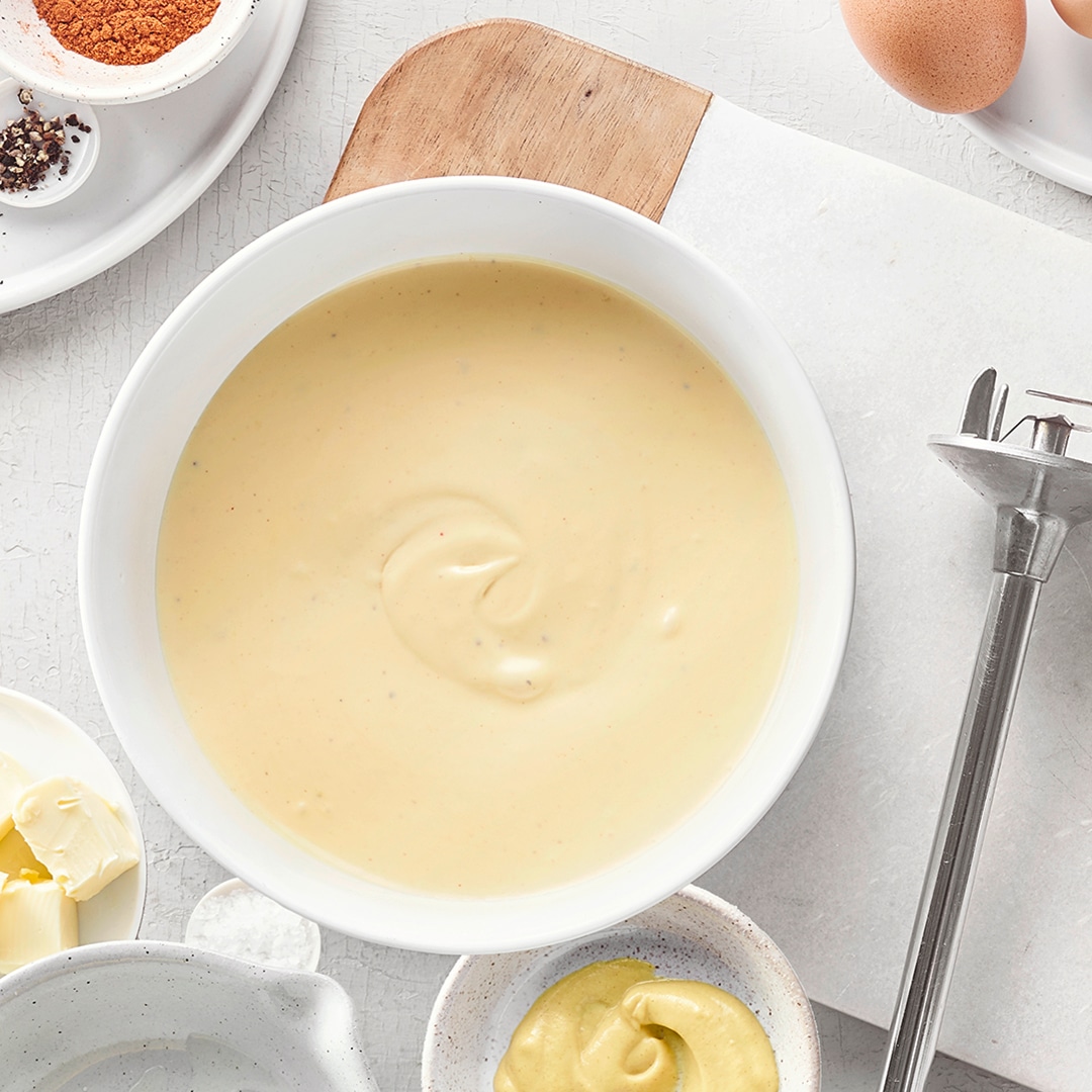 Schüssel mit cremiger Sauce, daneben Eier, Butter, Gewürze und ein Stabmixer auf heller Arbeitsfläche.