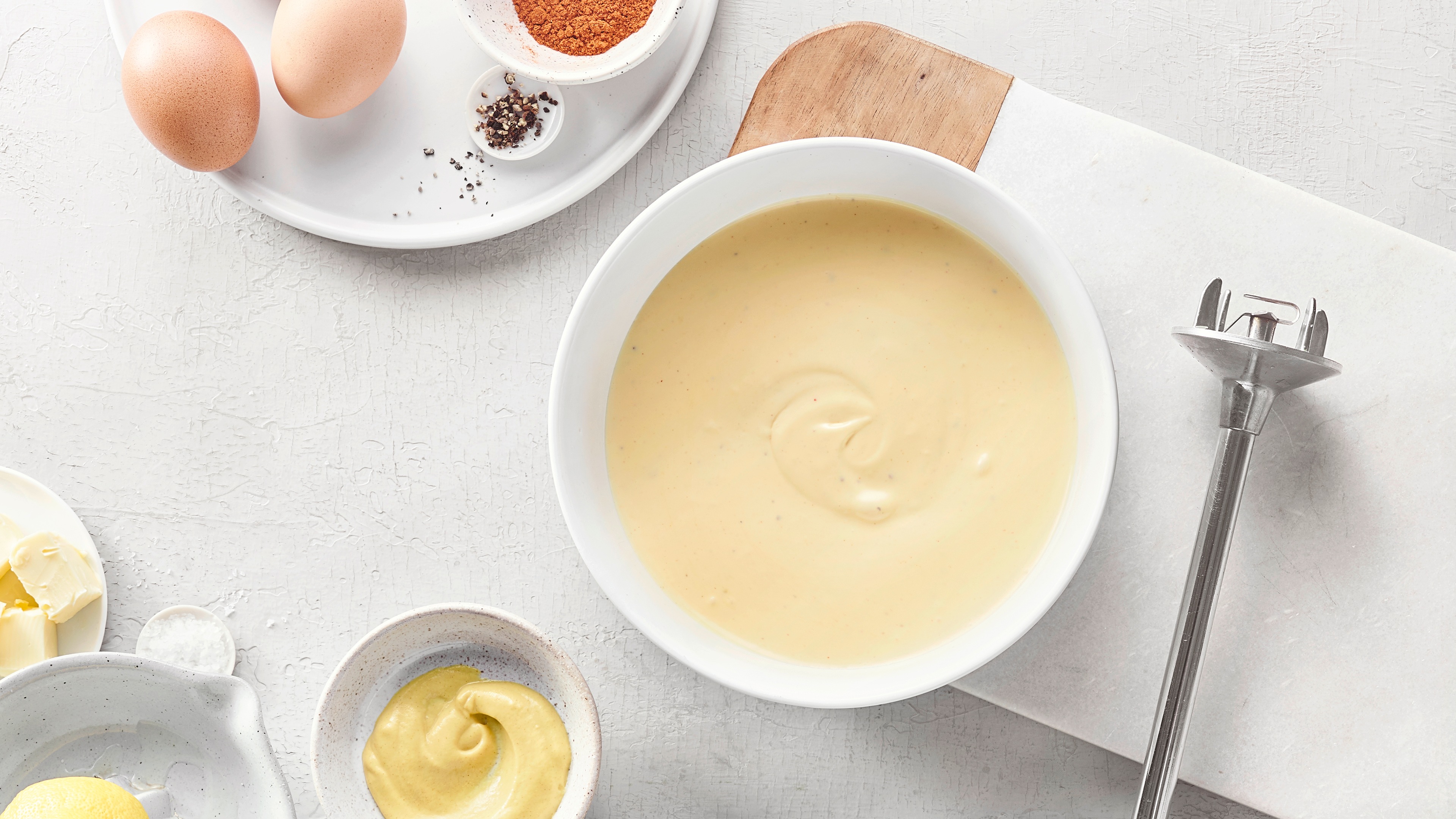 Schüssel mit cremiger Sauce, daneben Eier, Butter, Gewürze und ein Stabmixer auf heller Arbeitsfläche.