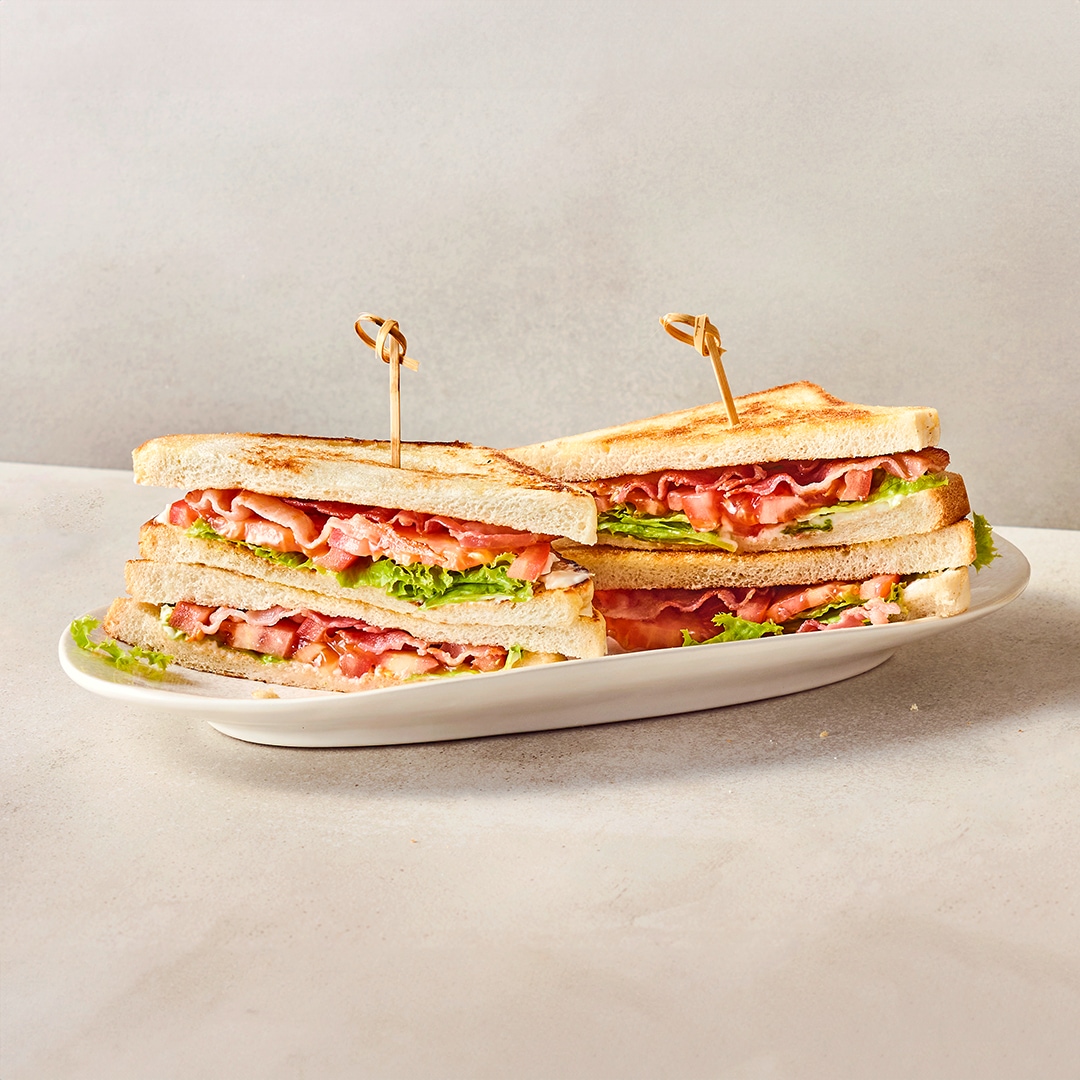 Ein ovaler Teller mit getoasteten Doppeldecker-Sandwiches, belegt mit Schinken, Tomatenscheiben und frischem Salat