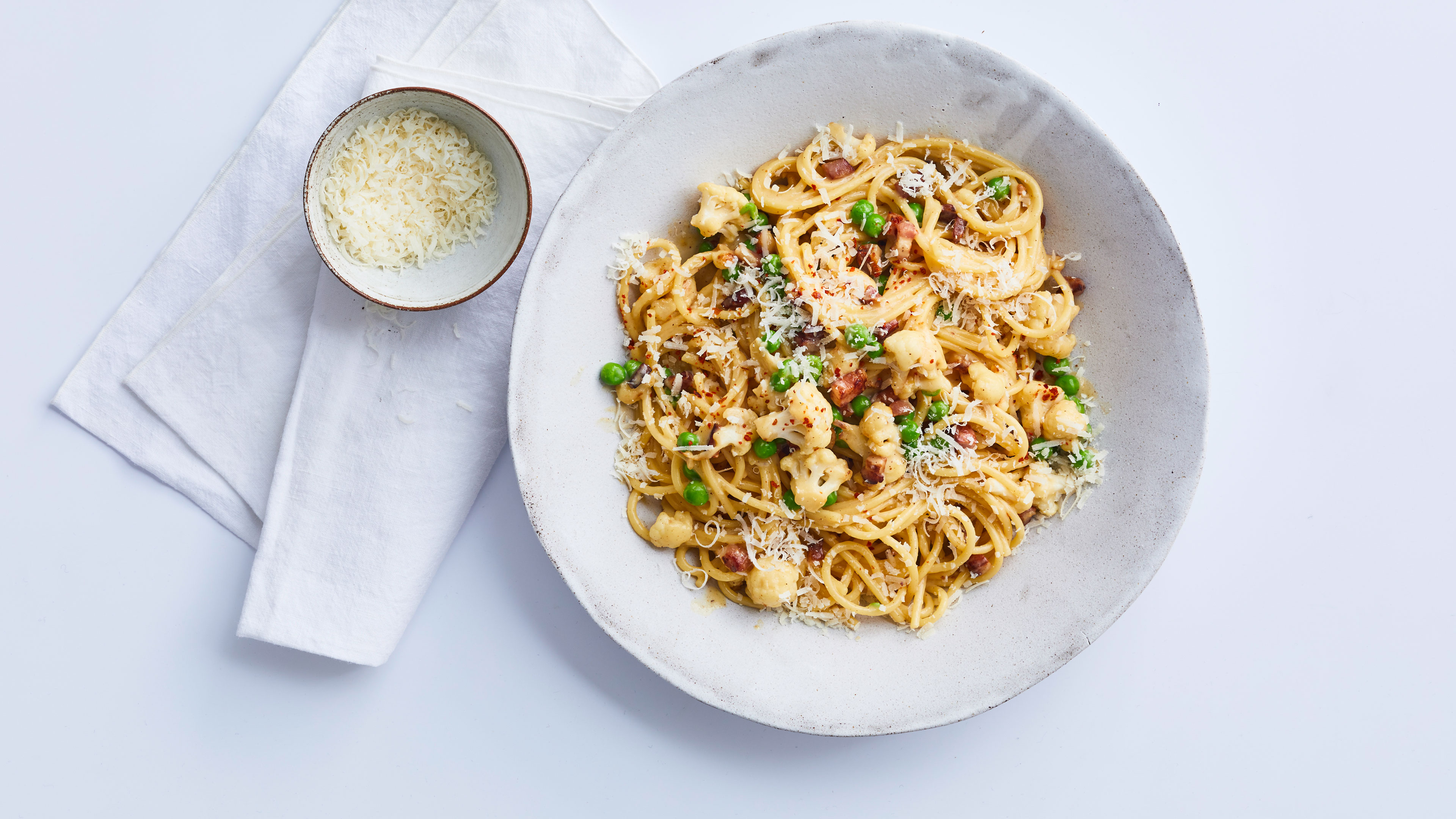 Teller mit Spaghetti, Blumenkohlröschen, Erbsen und Speckwürfeln, mit geriebenem Käse bestreut. Daneben steht eine kleine Schale mit weiterem geriebenem Käse auf einer weißen Serviette.