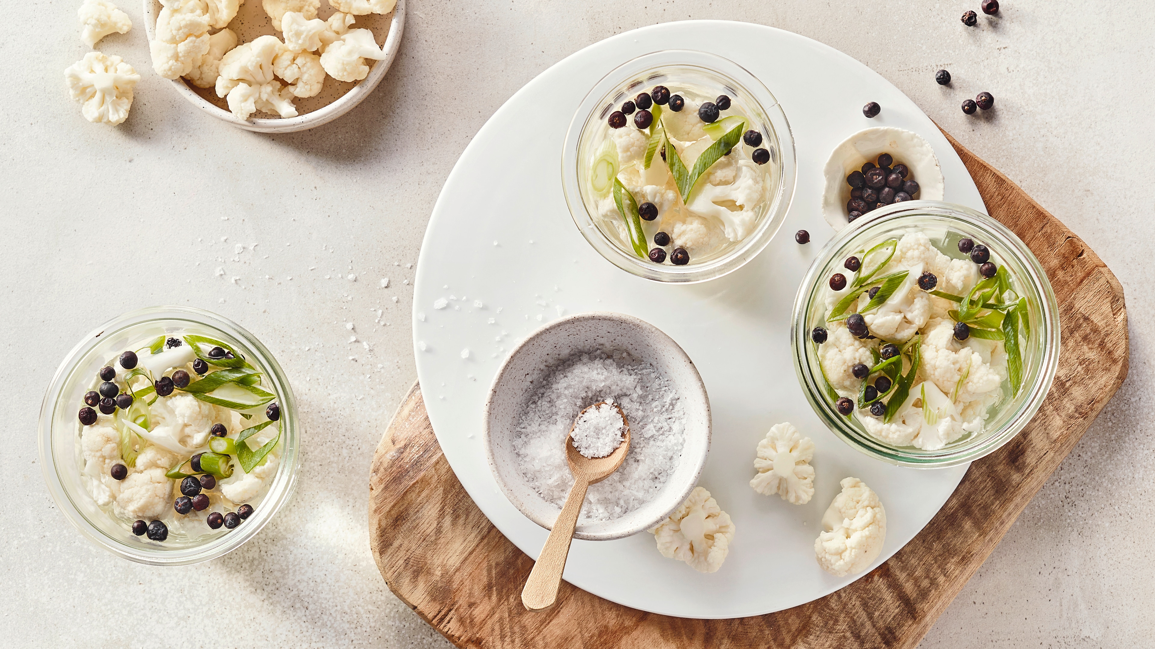 Ein Brett mit zwei Gläsern, gefüllt mit Blumenkohl, grünen Lauchstreifen und schwarzen Pfefferkörnern, daneben Salz.
